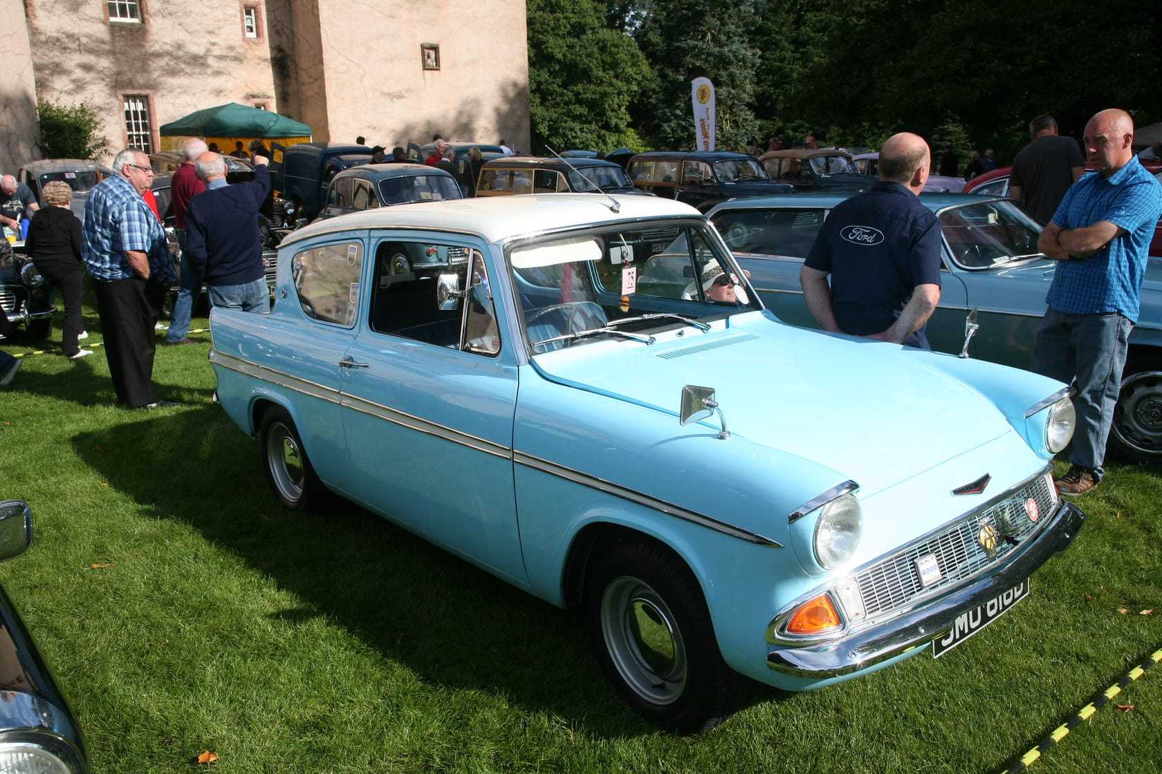 A pale blue Ford Anglia 105E Super at a classic-car show, front three-quarter view showing the reverse-rake rear window