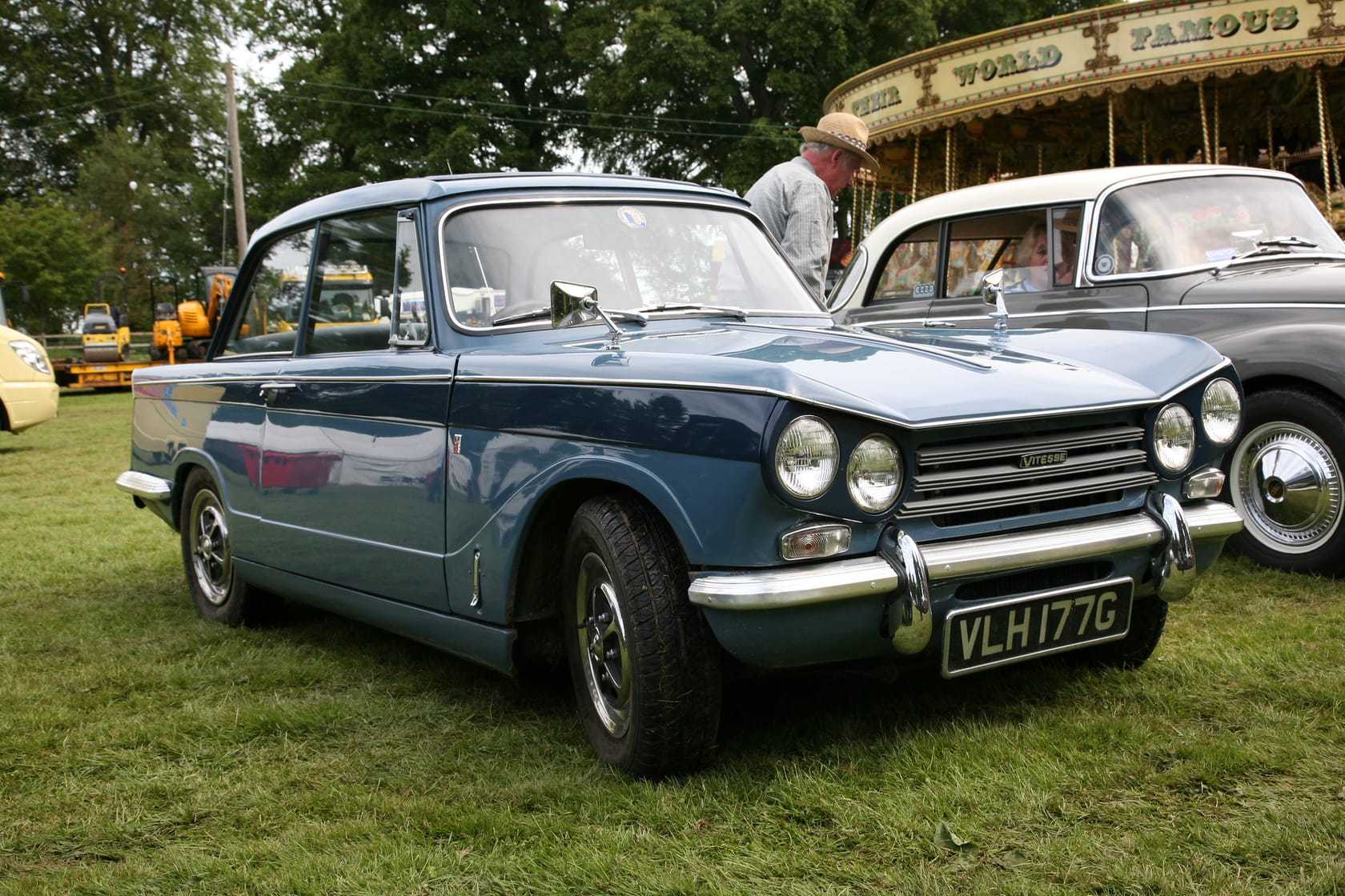 A blue and white 1969 Triumph Vitesse Mk2 saloon at a country fair, front three-quarter view, UK registration VLH 177G