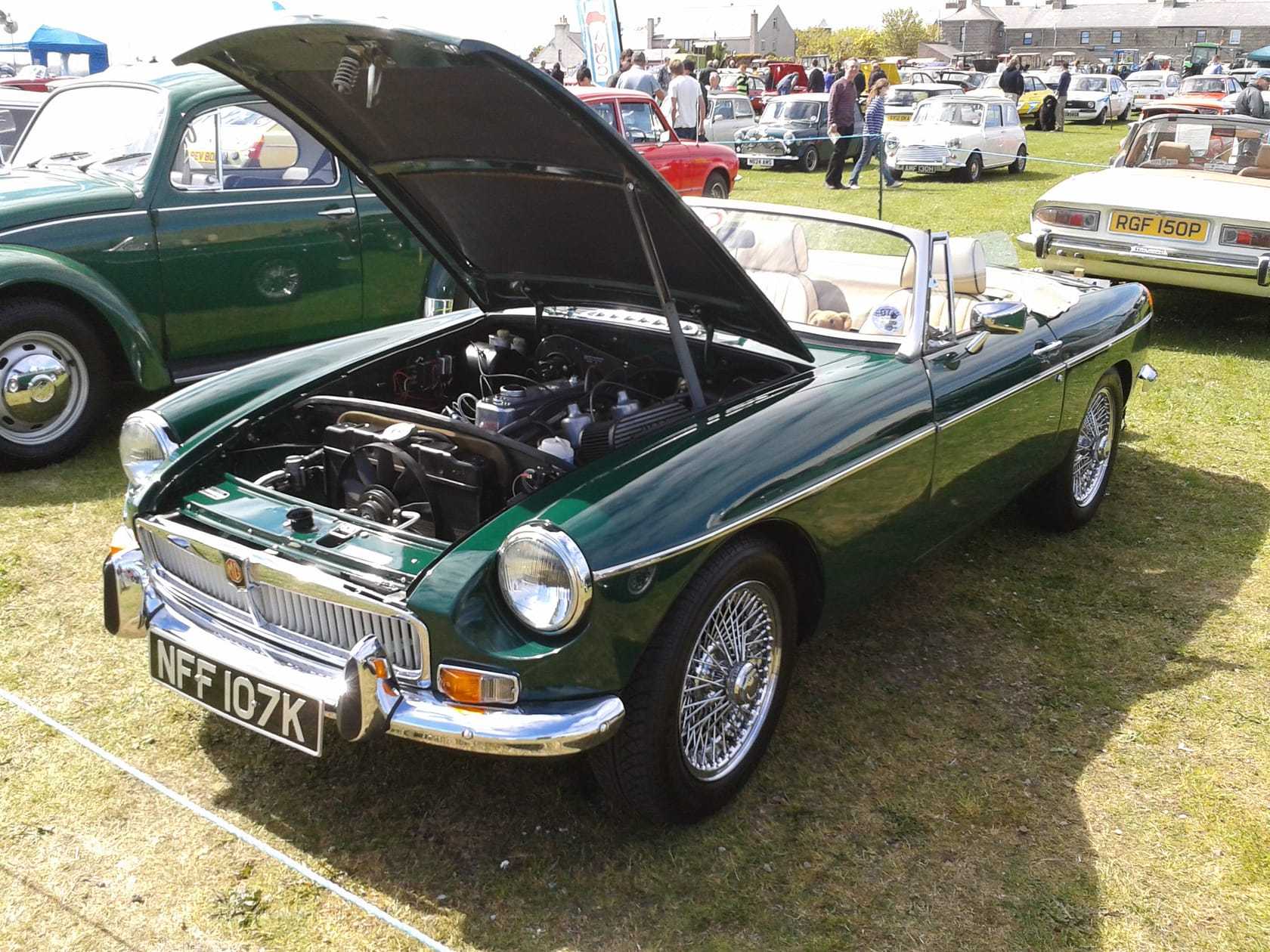A British Racing Green MGB roadster with its bonnet open at a classic car show surrounded by other classics