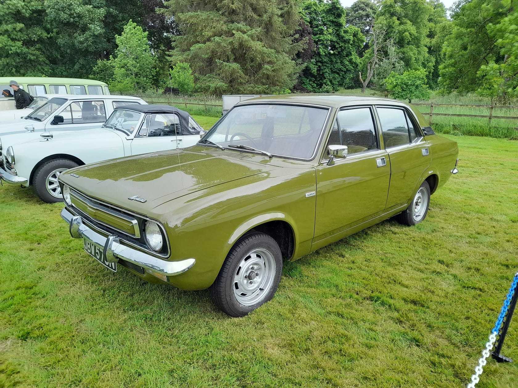 An olive green 1975 Morris Marina four-door saloon at an outdoor classic car show, front three-quarter view
