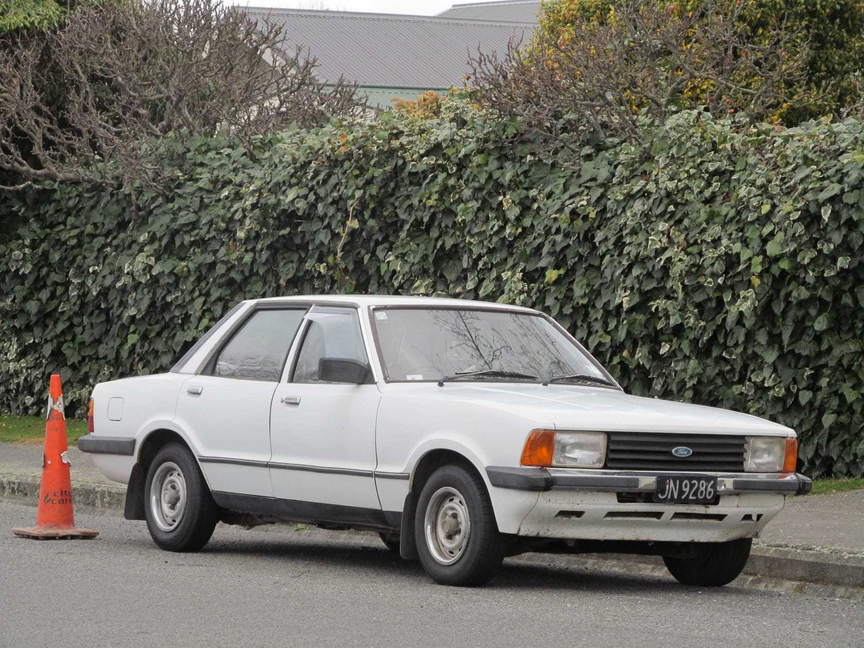A white 1980 Ford Cortina saloon parked beside a hedge, wearing a UK number plate
