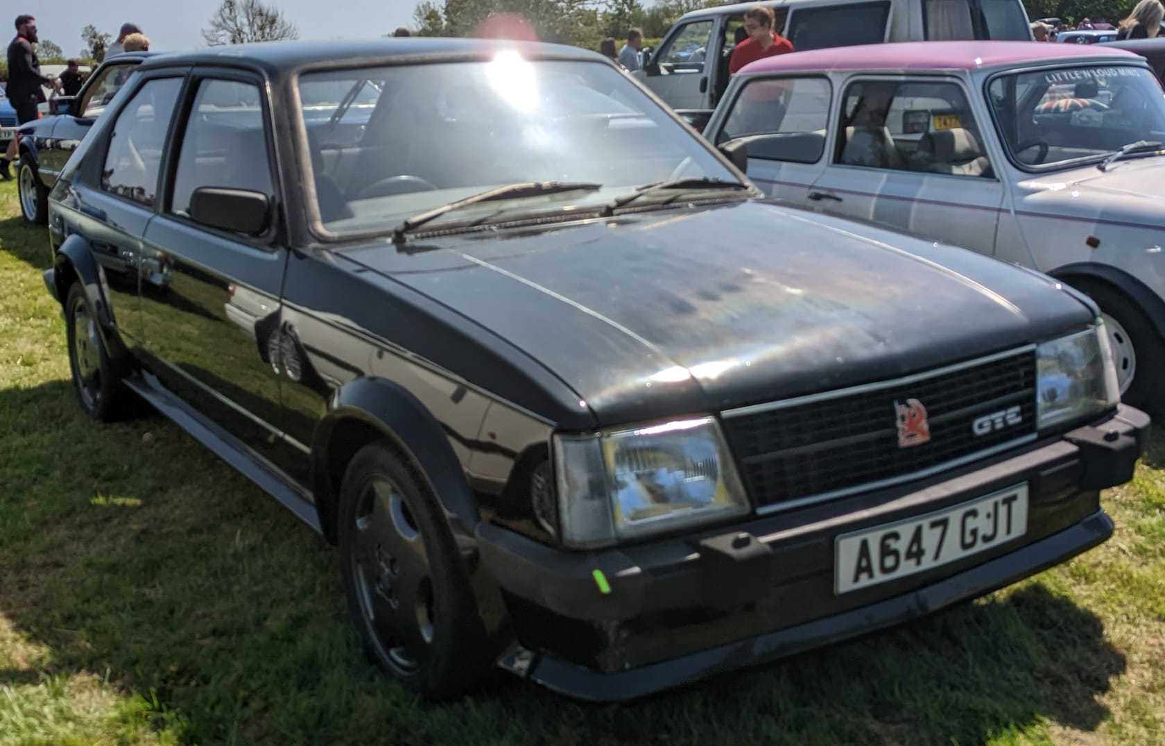 A red 1983 Vauxhall Astra GTE Mk1 with a UK registration on a show field, a Jaguar behind it