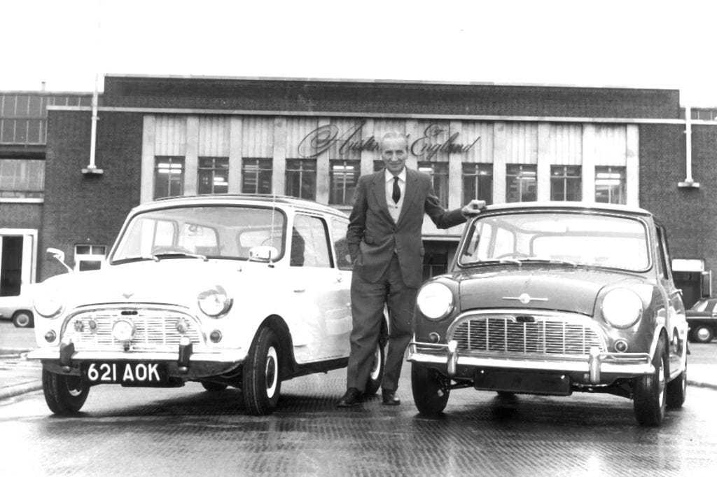 Alec Issigonis in a suit, standing between two early Minis outside the Austin works at Longbridge in 1965; the Mini on the left wears the registration 621 AOK
