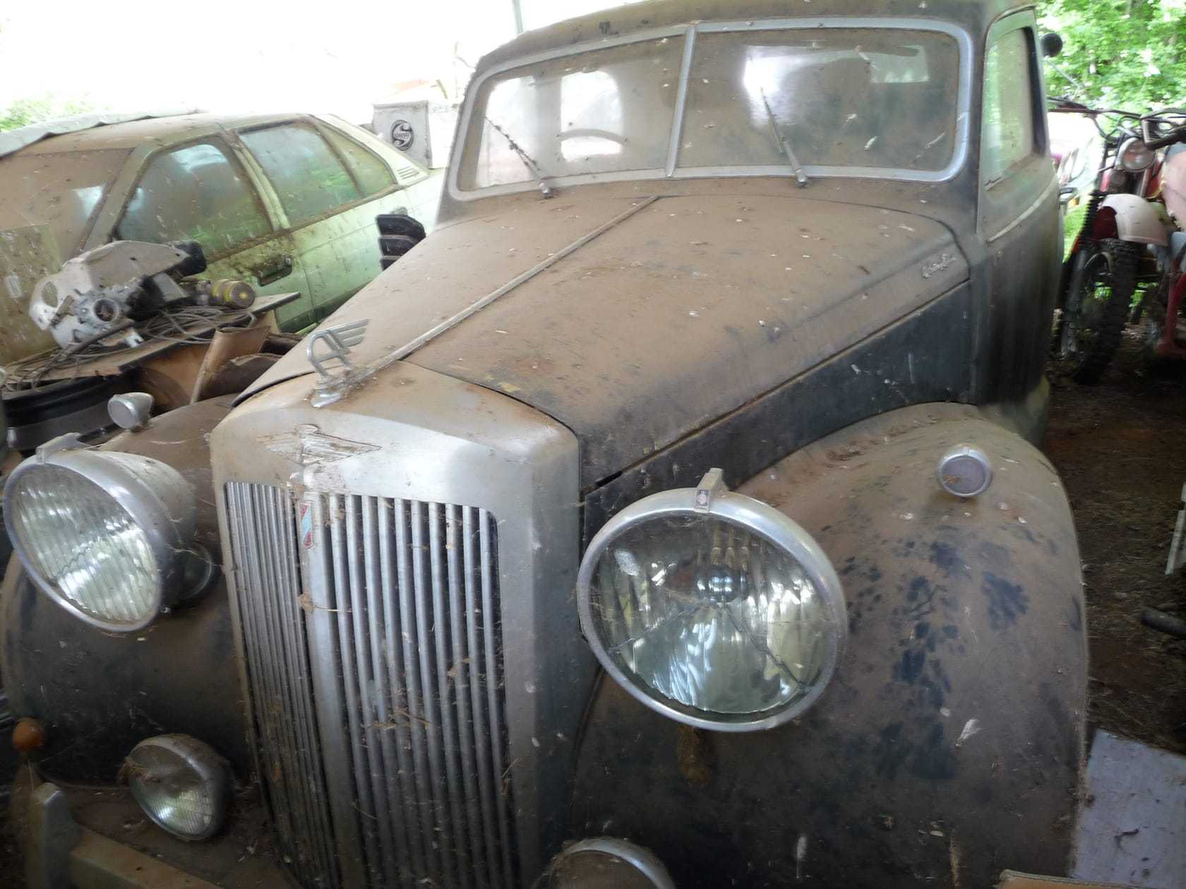 A dust-covered classic saloon with a chrome grille standing in a barn among other unrestored cars