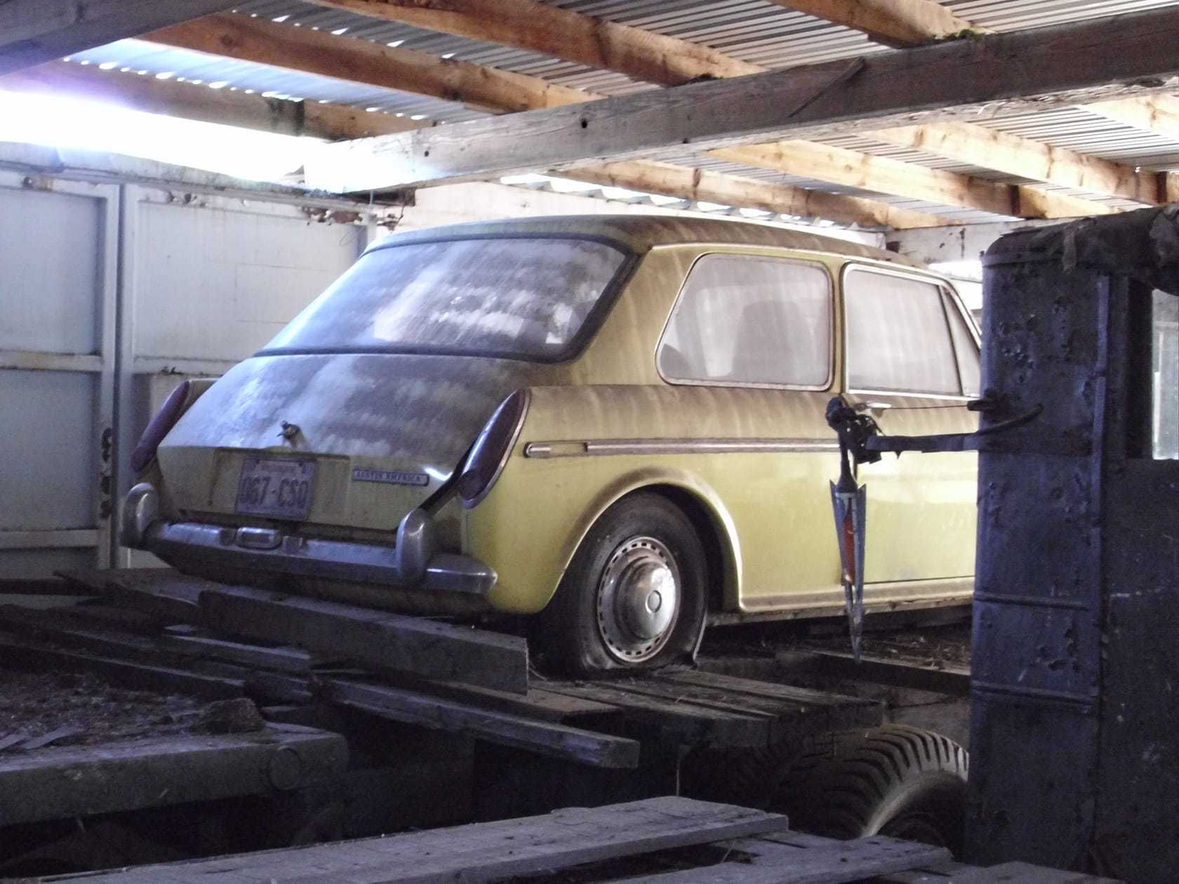 A dusty pale yellow Austin 1100 saloon stored on a raised wooden platform inside a barn