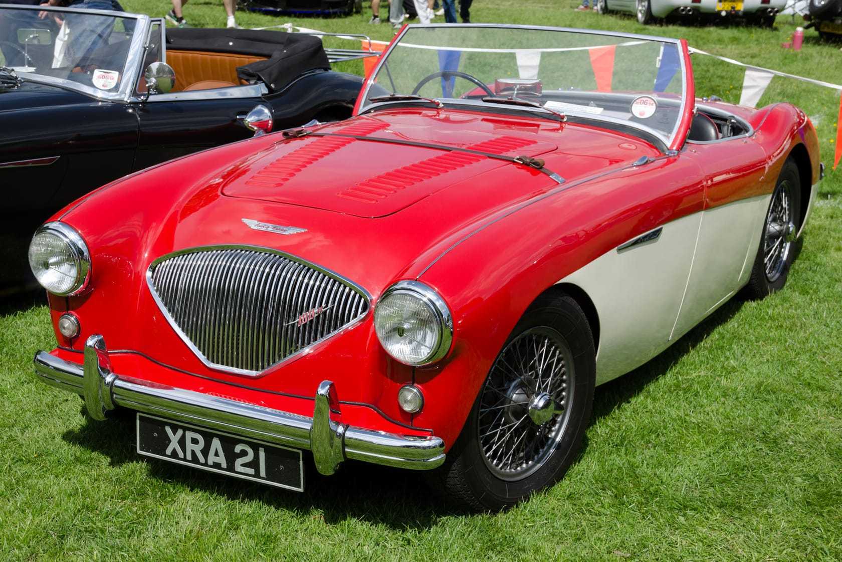A red and white 1955 Austin-Healey 100M with a louvred bonnet and a UK registration on a show field