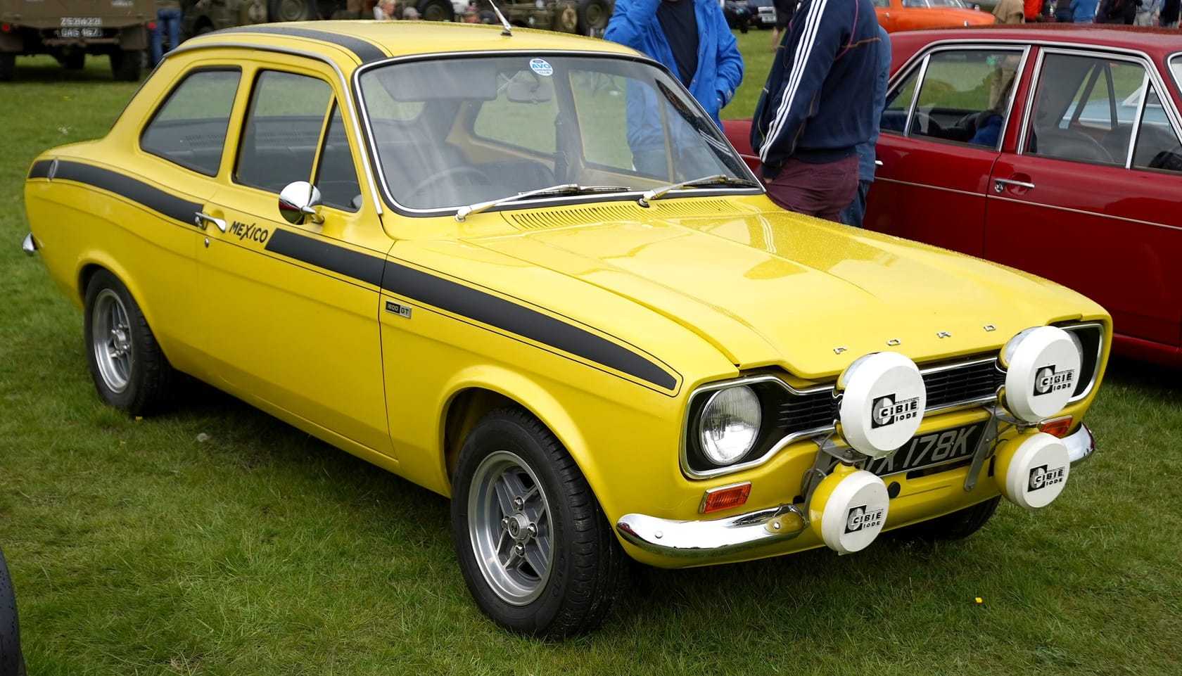 A yellow Mk1 Ford Escort Mexico with rally lamps and a Mexico side stripe, front three-quarter view