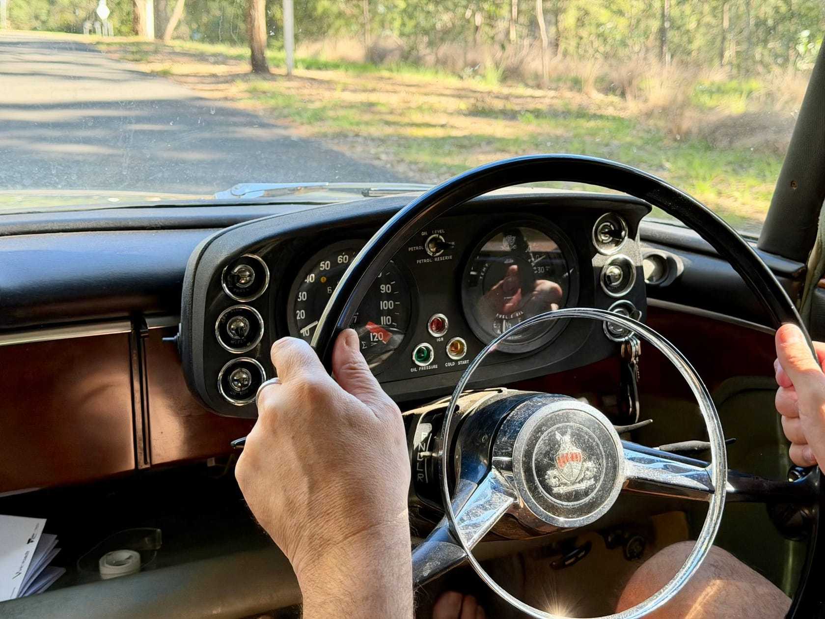 Close-up of a driver's hands on the thin steering wheel of a classic car, with the road ahead through the windscreen