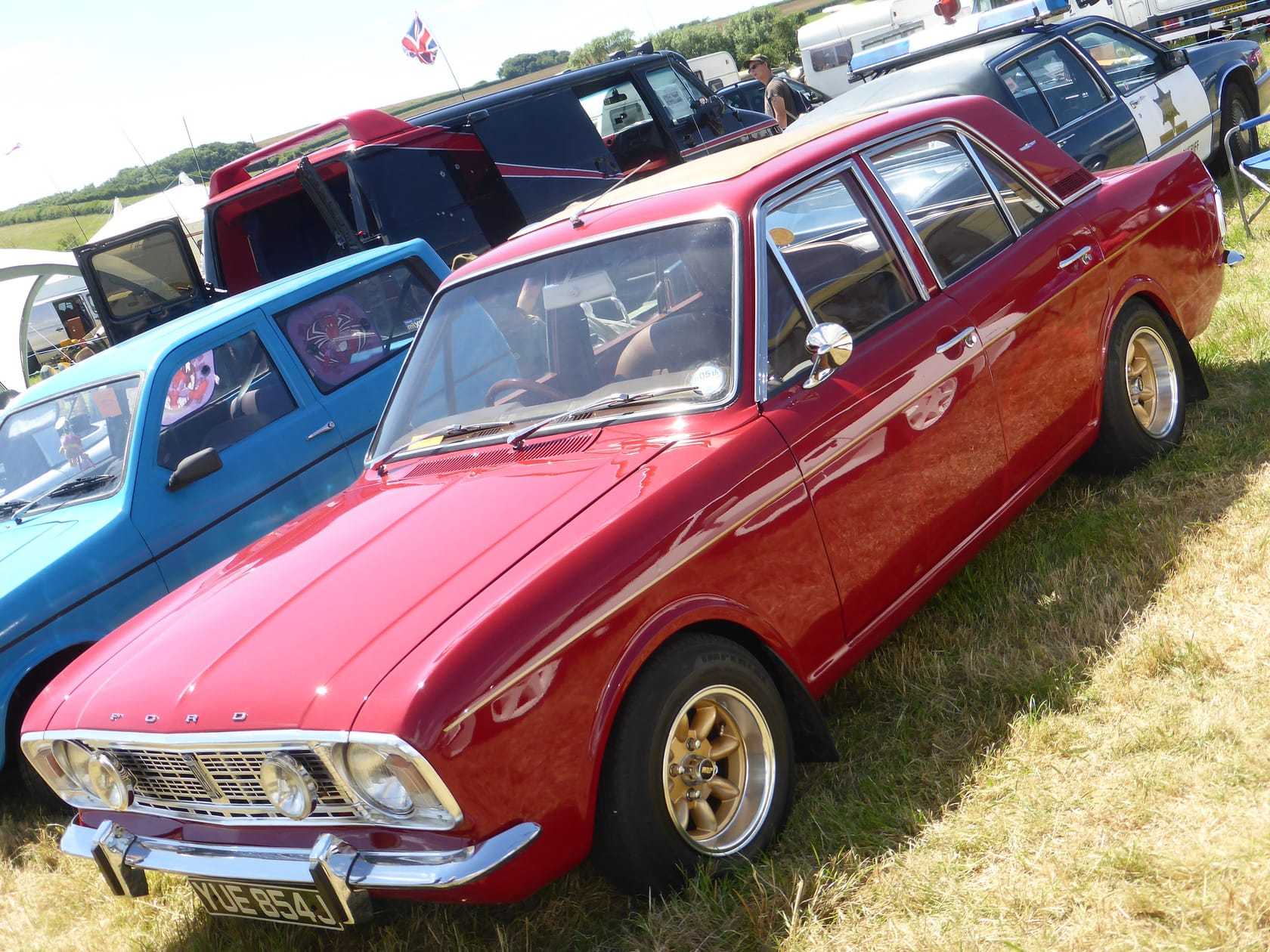 A red 1970 Ford Cortina 1600E with gold Rostyle wheels at a classic car show, front three-quarter view