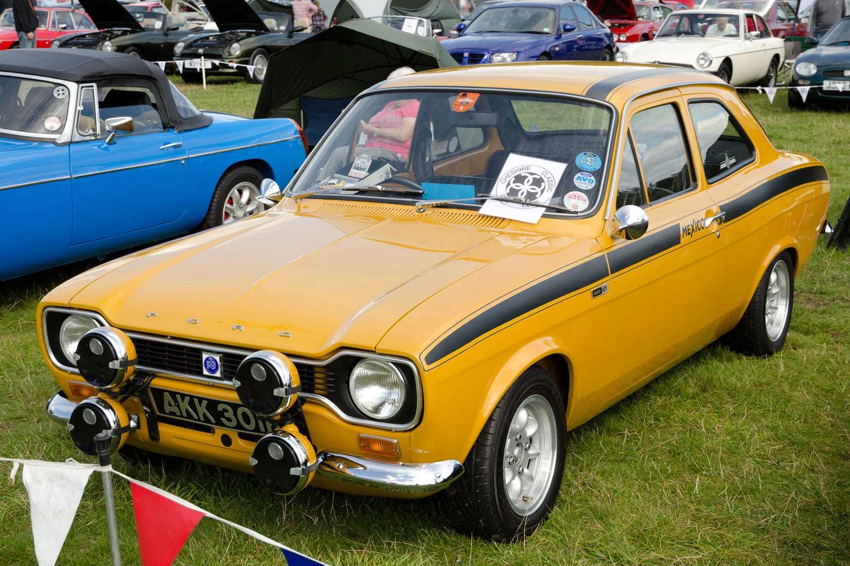 A yellow 1971 Ford Escort Mk1 Mexico with a black side stripe and rally lamps, UK registration, on a show field