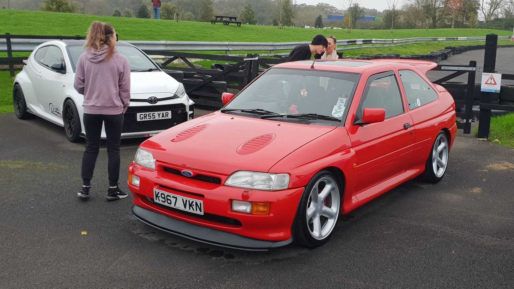 A red Ford Escort RS Cosworth with bonnet vents and a front splitter, front three-quarter view at a track meeting