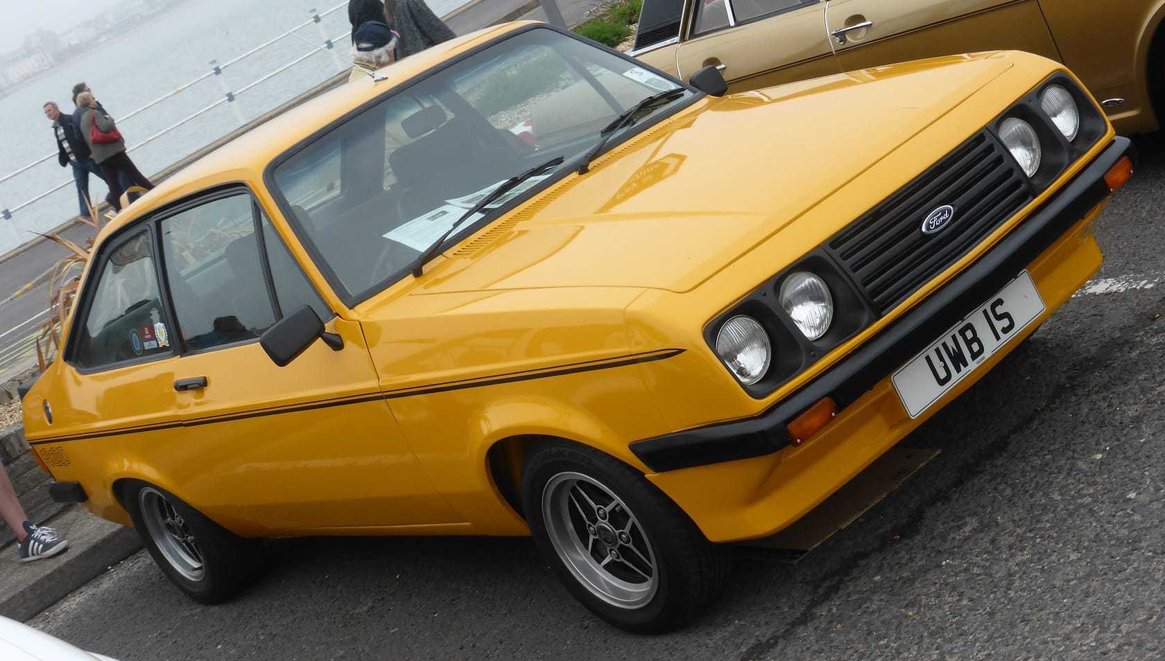 A yellow Ford Escort Mk2 RS2000 with its black polyurethane droop-snoot nose, front three-quarter view at a seafront classic car gathering