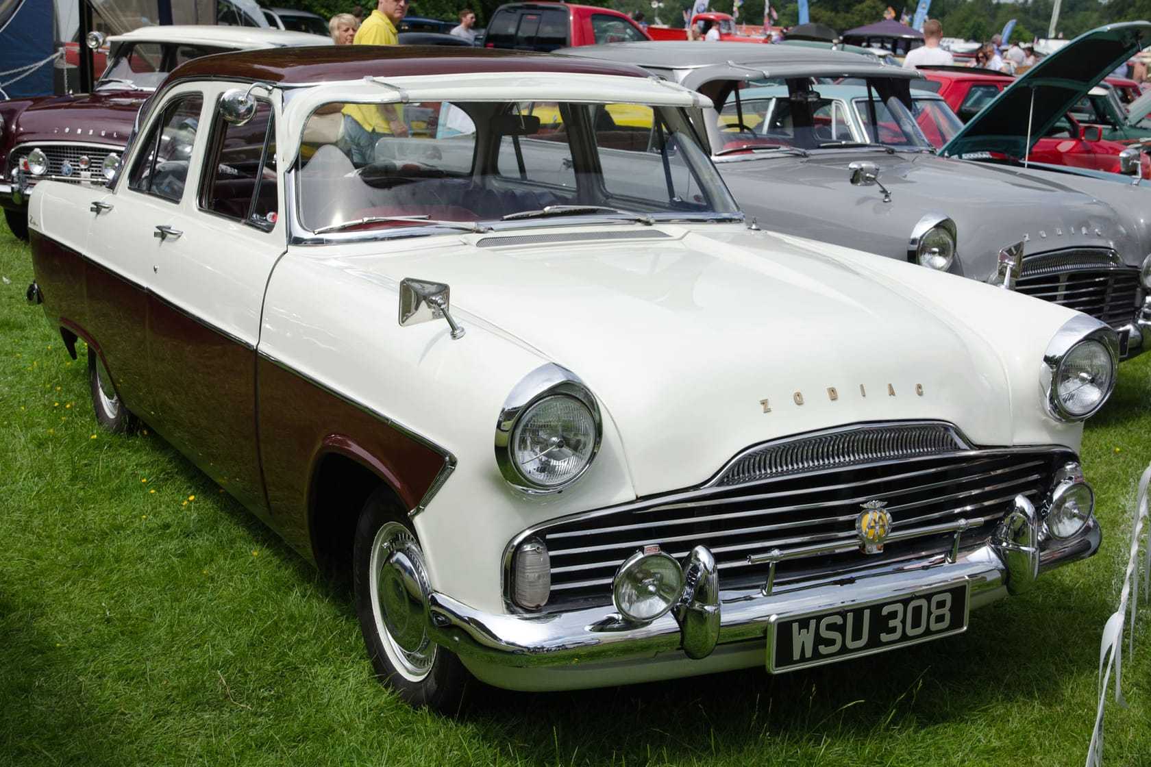 A two-tone cream-and-maroon Ford Zodiac Mk2 with tail fins, front three-quarter view at a show