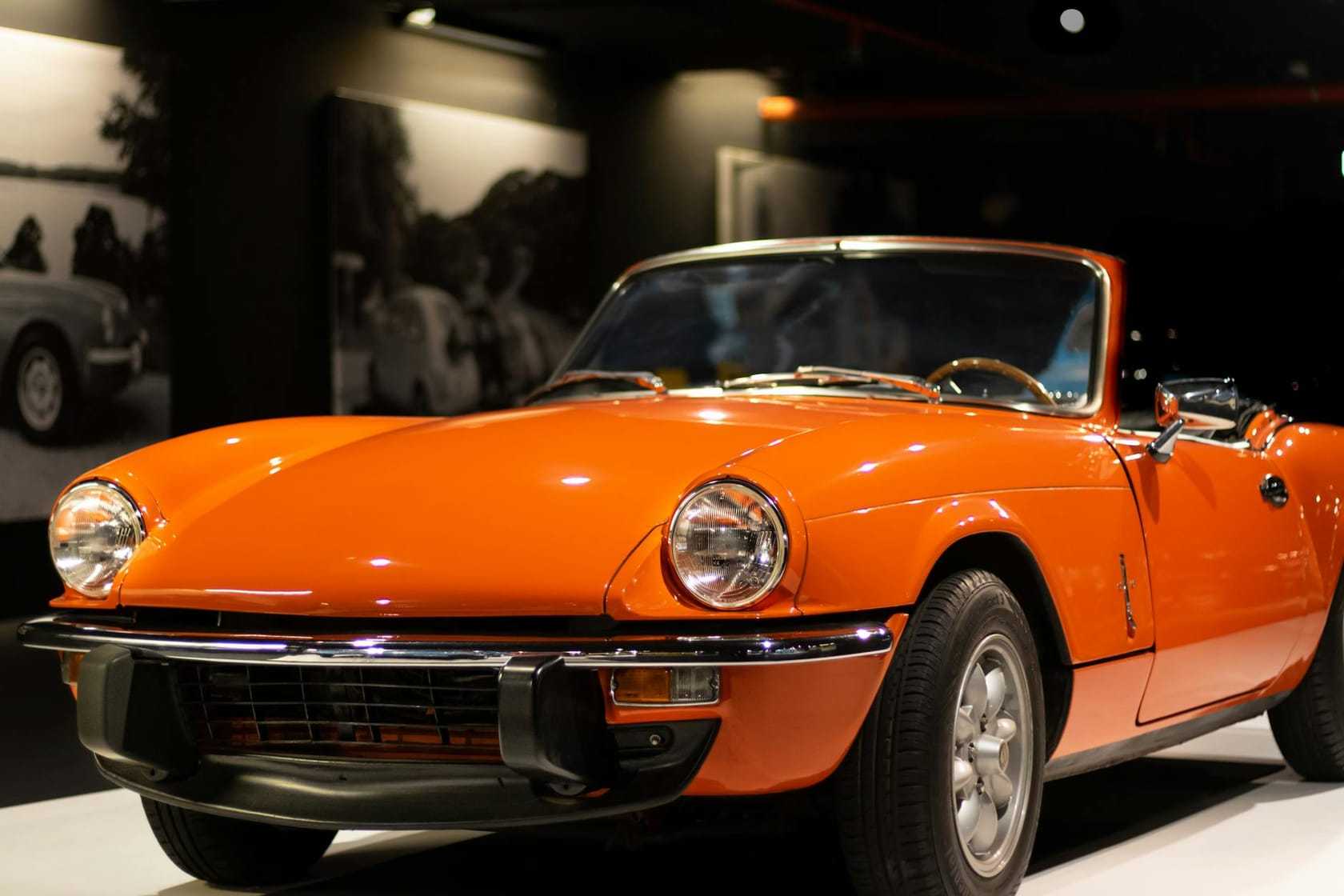 An orange Triumph Spitfire convertible, top down, front three-quarter view on a museum display stand
