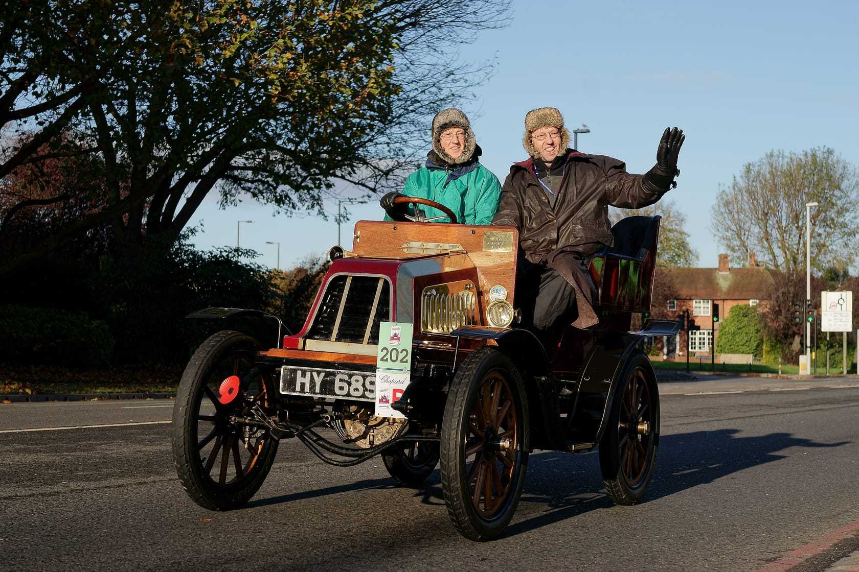 A veteran car from the turn of the twentieth century with two occupants in winter coats, one waving, on a tree-lined suburban road during the London to Brighton Veteran Car Run