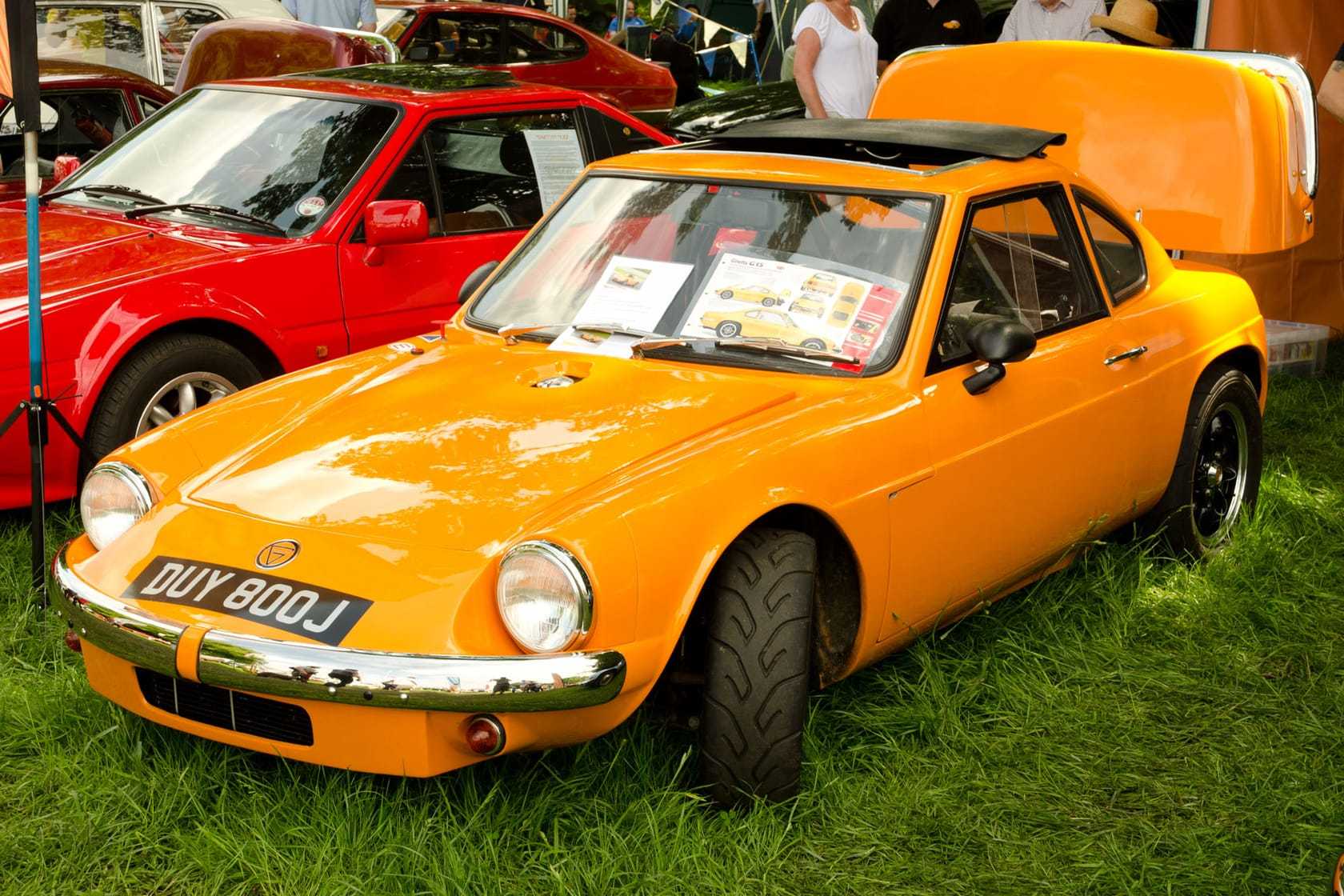 An orange Ginetta G15 coupe with a black roof panel, registration DUY 800J, three-quarter front view at an outdoor classic car show