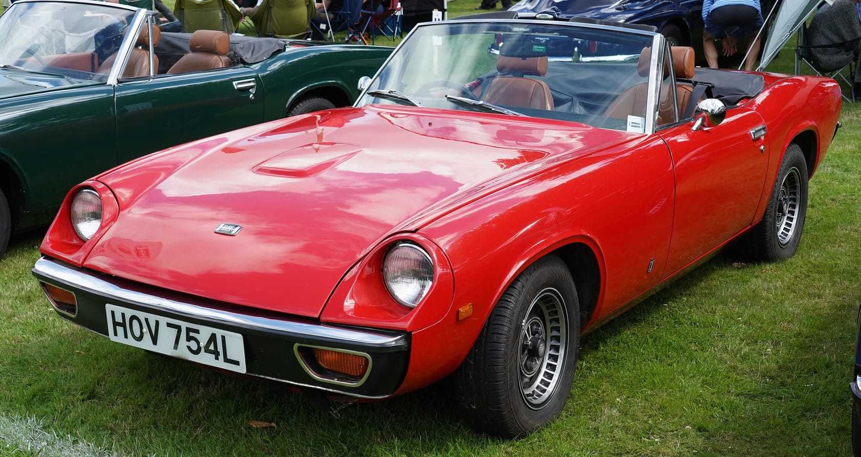 A red Jensen-Healey roadster with the top down at a show, front three-quarter view, UK registration HOV 754L