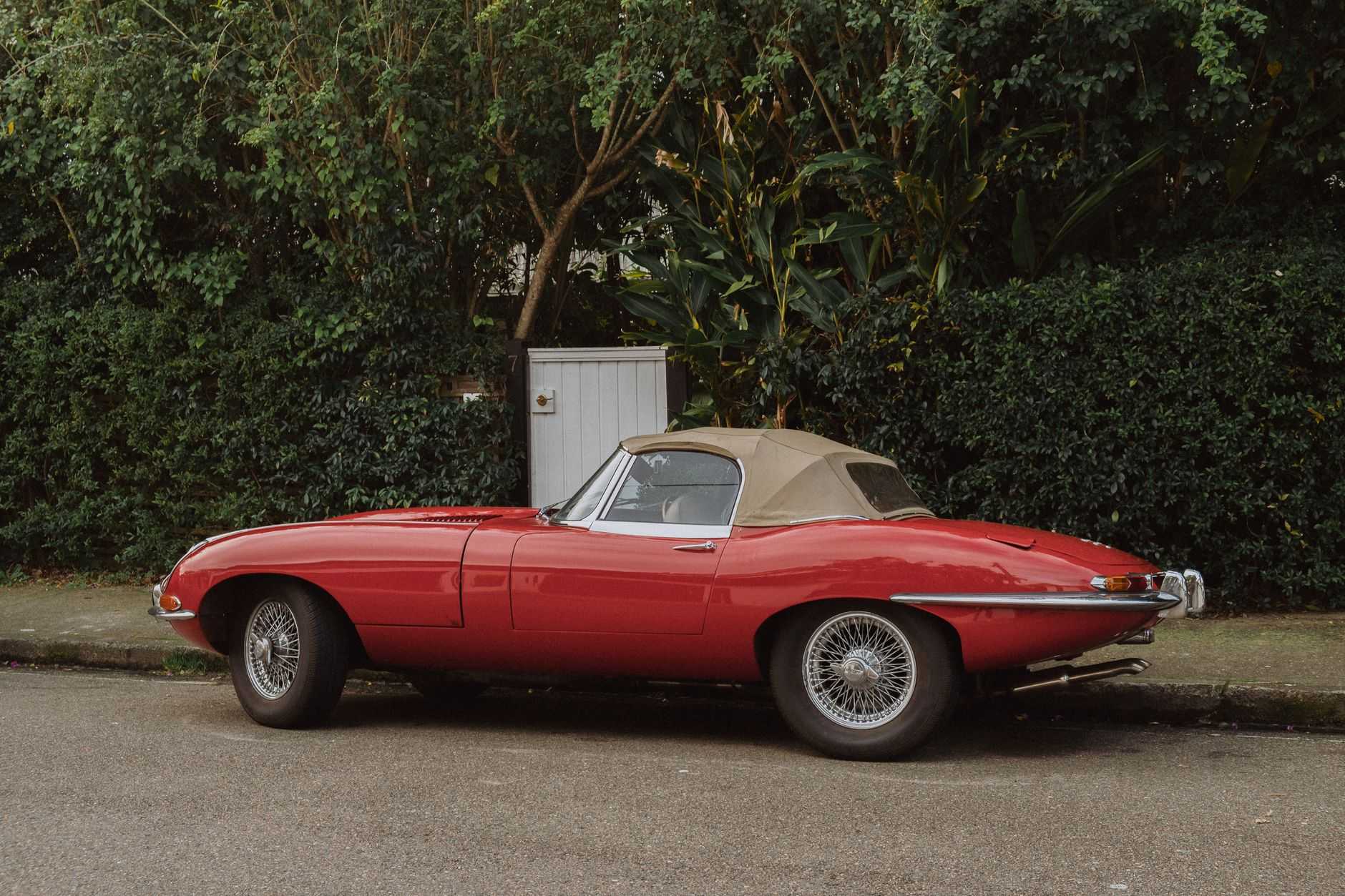 A red Jaguar E-Type roadster with the soft top raised and chrome wire wheels, parked in profile beside a green hedge