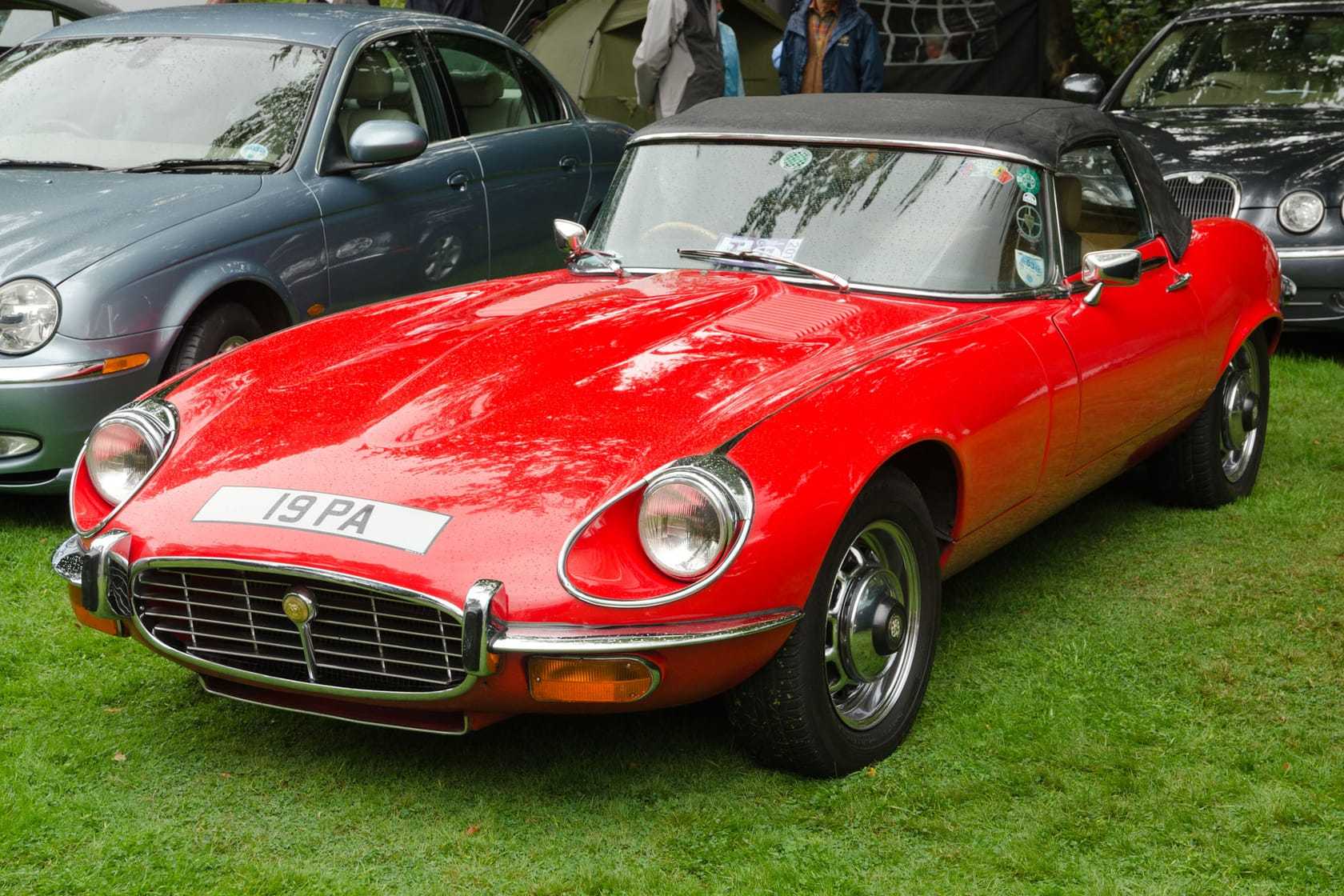 A red 1975 Jaguar E-Type Series 3 V12 roadster with UK registration 19 PA, front three-quarter view showing the cross-slat grille and flared arches