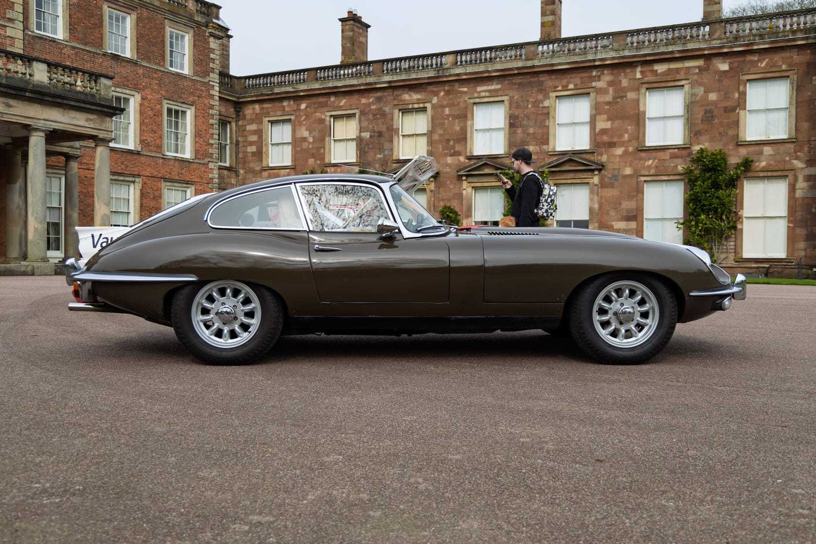 A grey Jaguar E-Type fixed-head coupe in side profile on the gravel in front of a country house