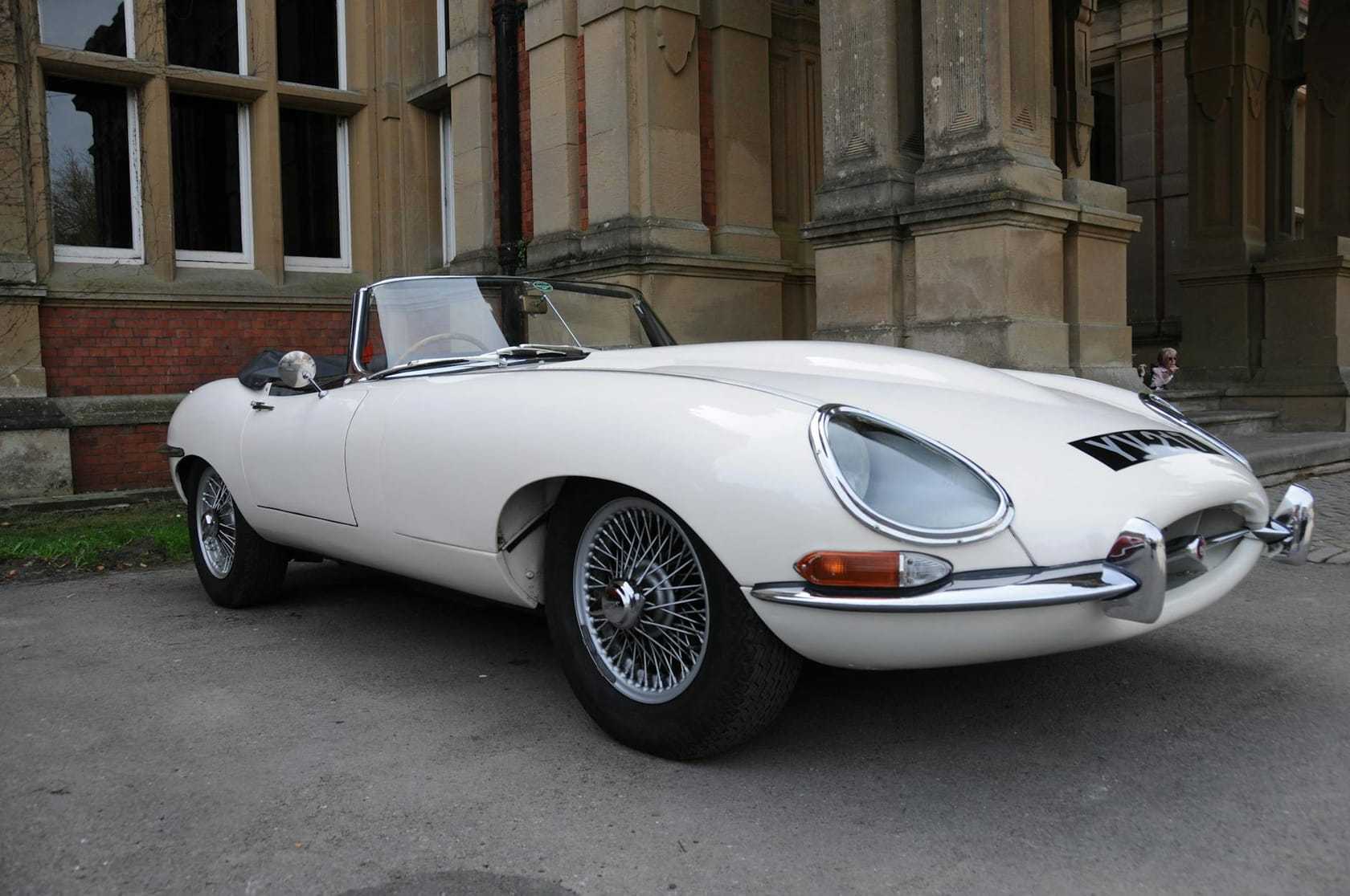 A white Jaguar E-Type Series 1 roadster with wire wheels parked beside a historic stone building, front three-quarter view