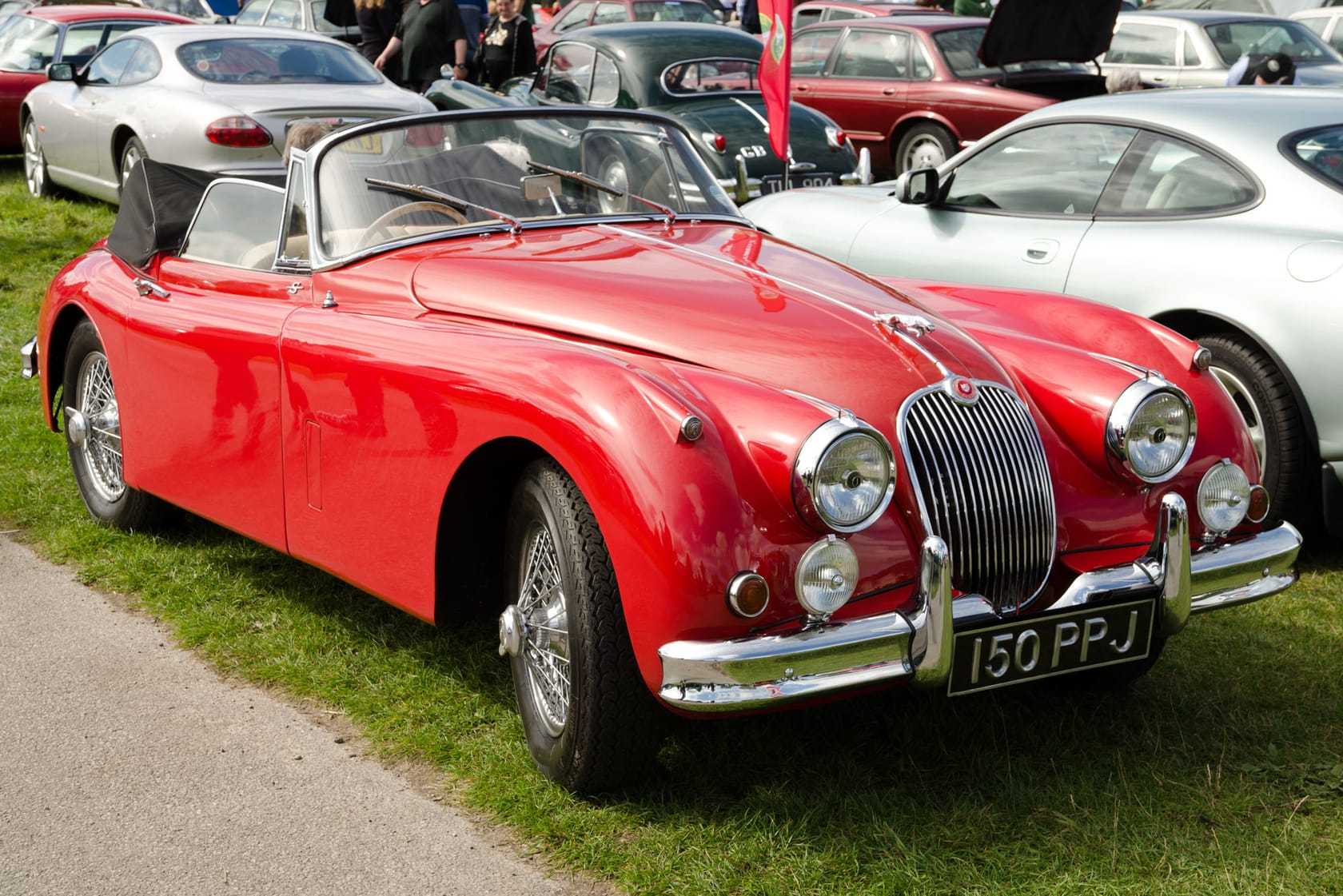 A red 1960 Jaguar XK150 roadster with a UK registration on a show field, its hood down