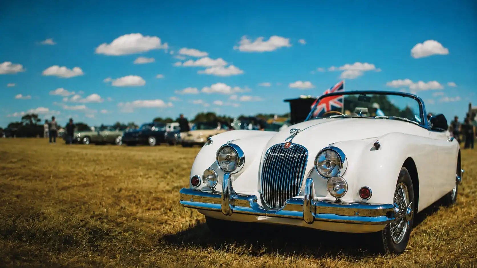 A white Jaguar XK150 drophead coupe at a British classic car show, a Union Jack flying behind the windscreen, front three-quarter view