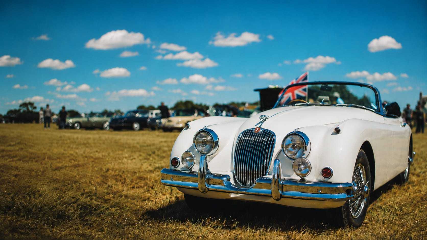 A white Jaguar XK150 drophead coupe at a British classic car show, a Union Jack flying behind the windscreen, front three-quarter view.