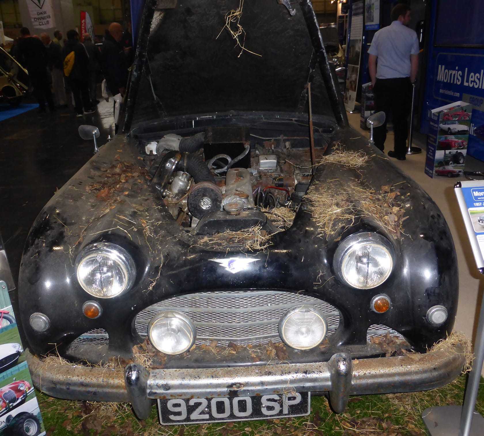 A dust and straw covered black Jensen saloon with its bonnet open, displayed at a show as an unrestored barn find