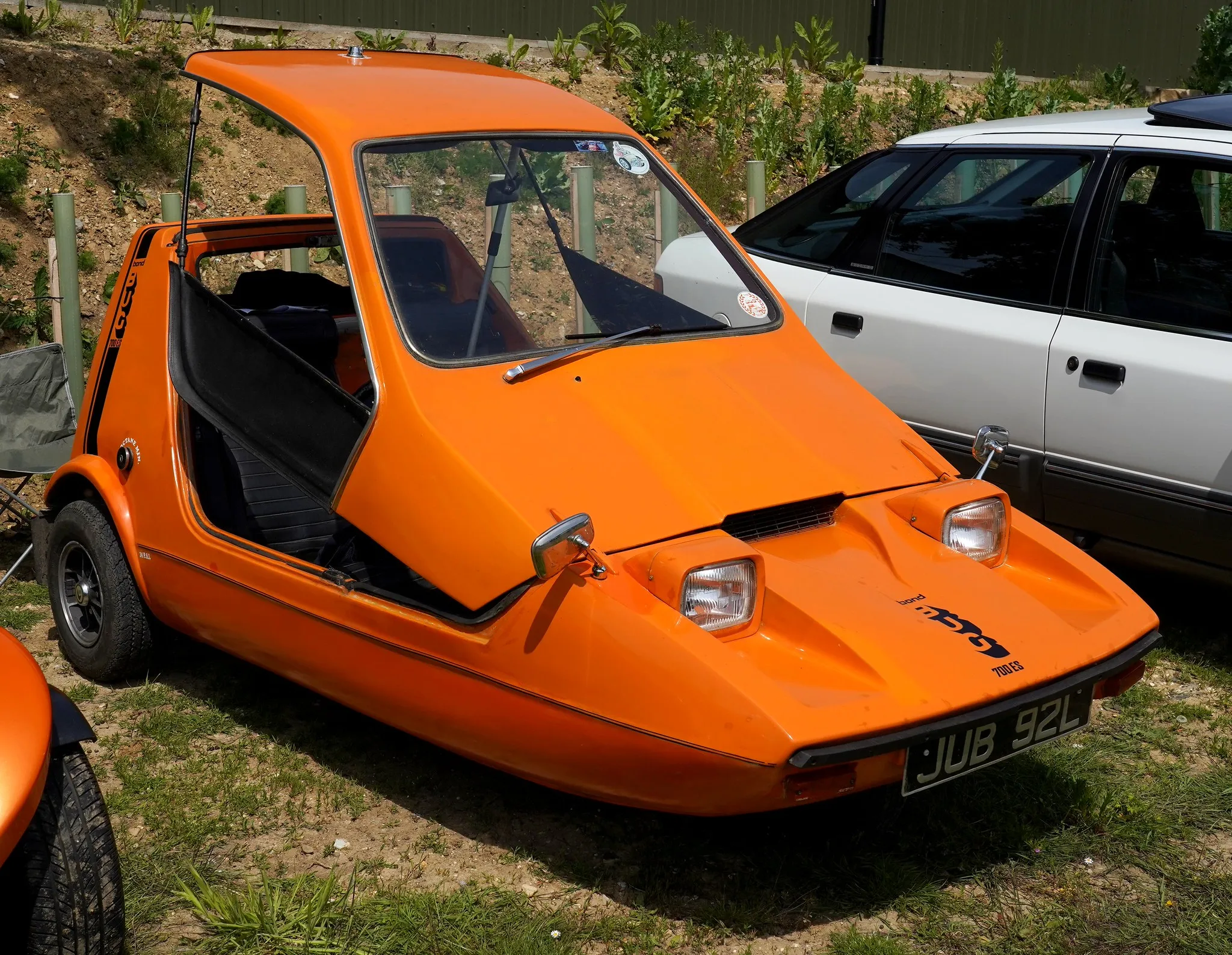 An orange Bond Bug three-wheeler at a show, front three-quarter view showing the wedge profile and the open doorless side with the seats visible