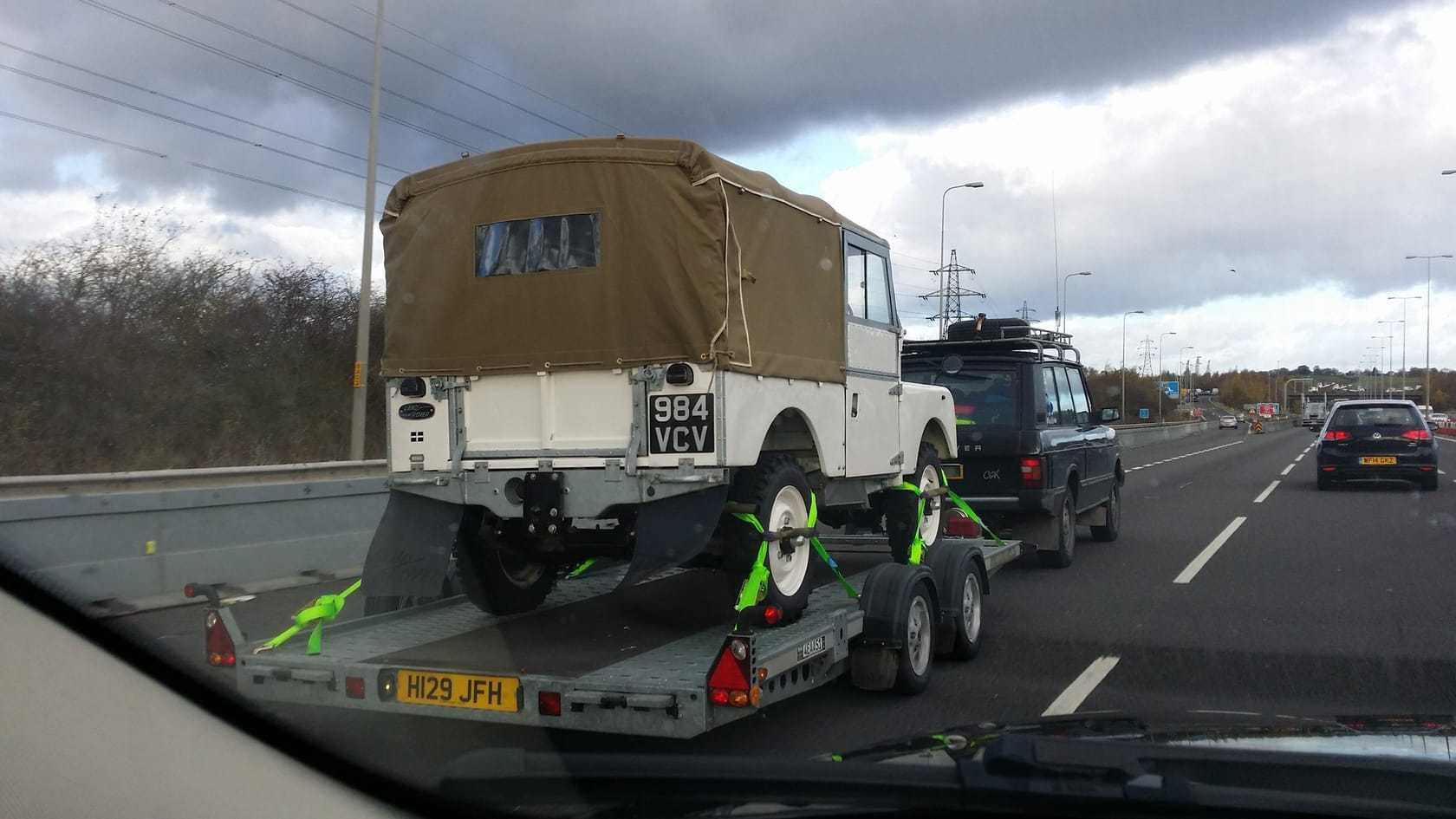 A cream 1955 Land Rover Series I with a canvas tilt strapped down on a car transporter trailer on a dual carriageway