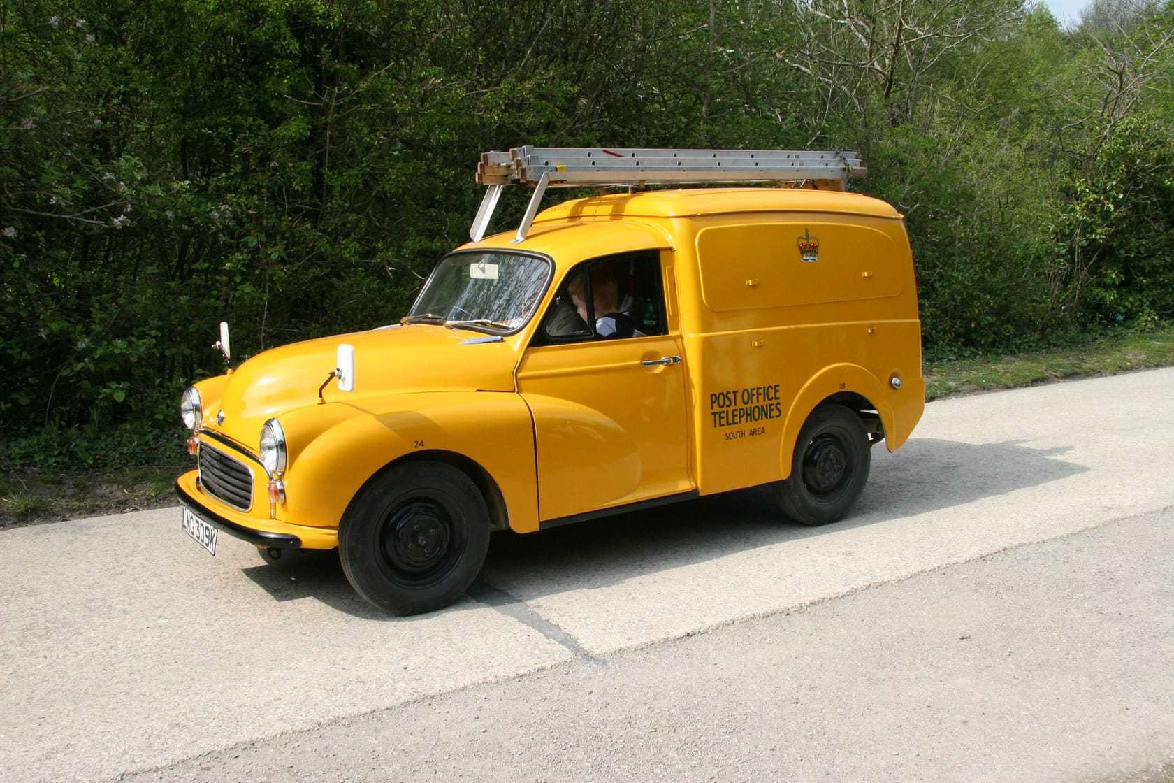 A yellow Post Office Telephones Morris Minor van with a ladder on the roof rack, on a country road