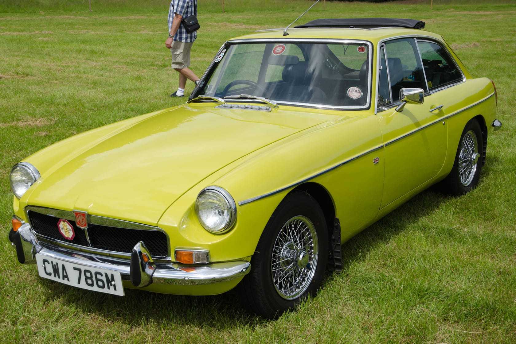 A yellow 1974 MGB GT with chrome wire wheels, a sunroof and a UK registration, parked on grass at a show