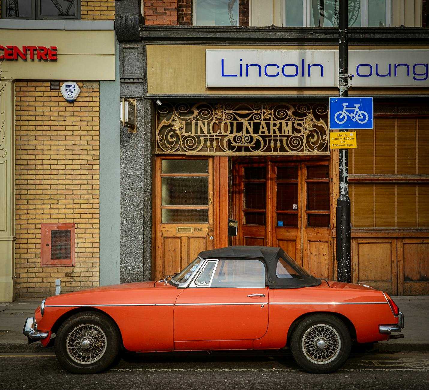 An orange MG MGB roadster with chrome wire wheels, side view parked outside a town pub