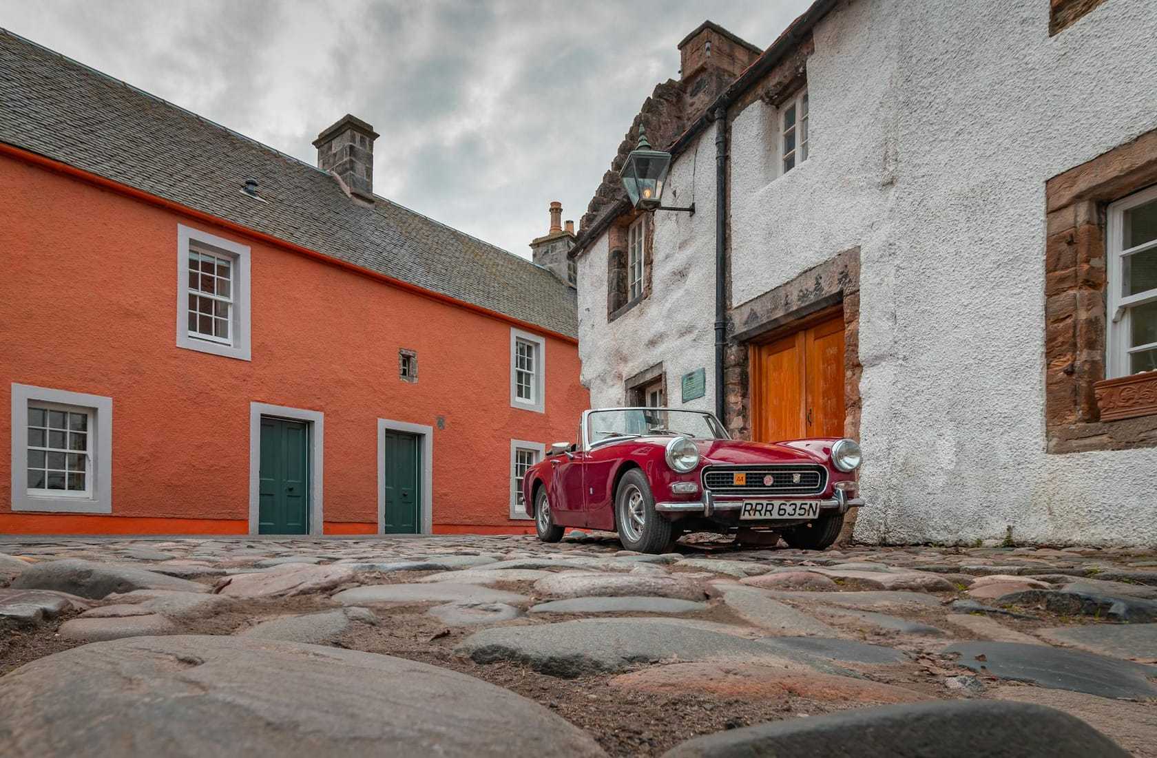 A dark red MG Midget with the top down, parked on a cobbled square between old stone buildings