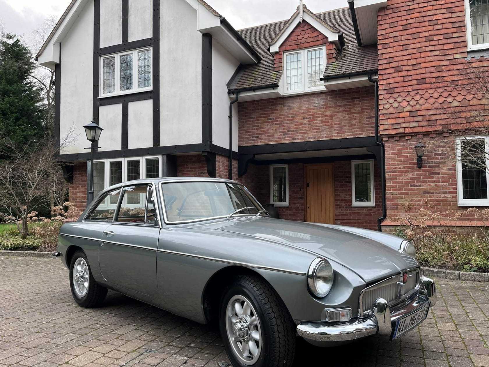 A silver MG MGB GT fastback coupe, front three-quarter view on a driveway in front of a mock-Tudor house