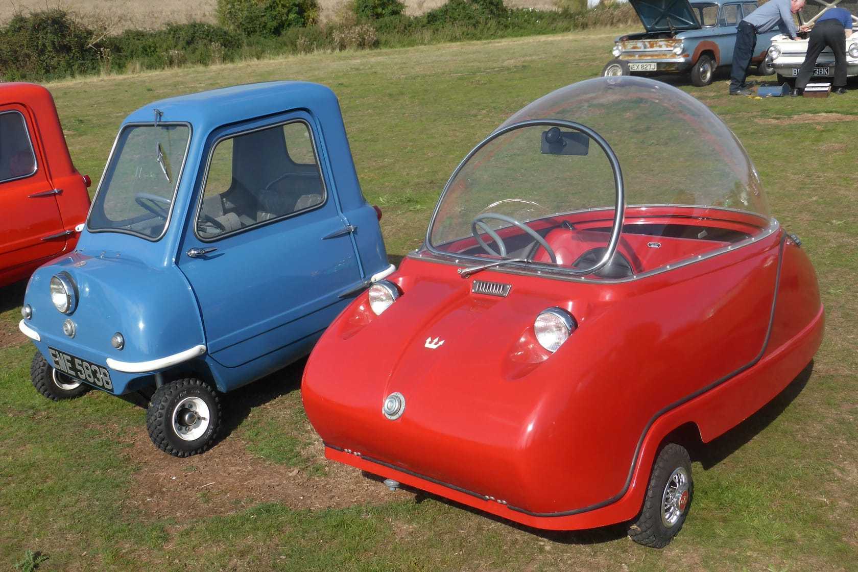 A blue Peel P50 and a red Peel Trident microcar side by side on grass at a show