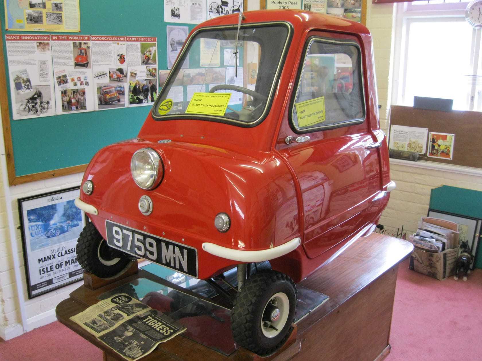 A red Peel P50 microcar on a display plinth at the Manx Transport Museum in Peel, Isle of Man, front three-quarter view showing the single door and three wheels
