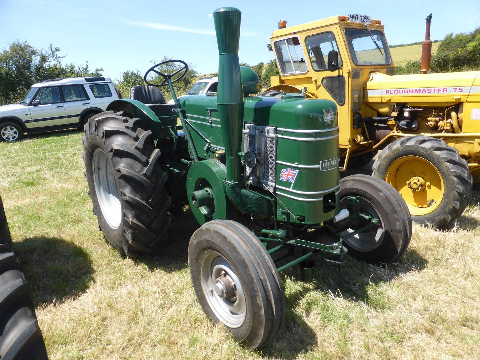 A green 1950s Field Marshall tractor with its tall single-cylinder exhaust, on a showground beside a yellow Ploughmaster tractor