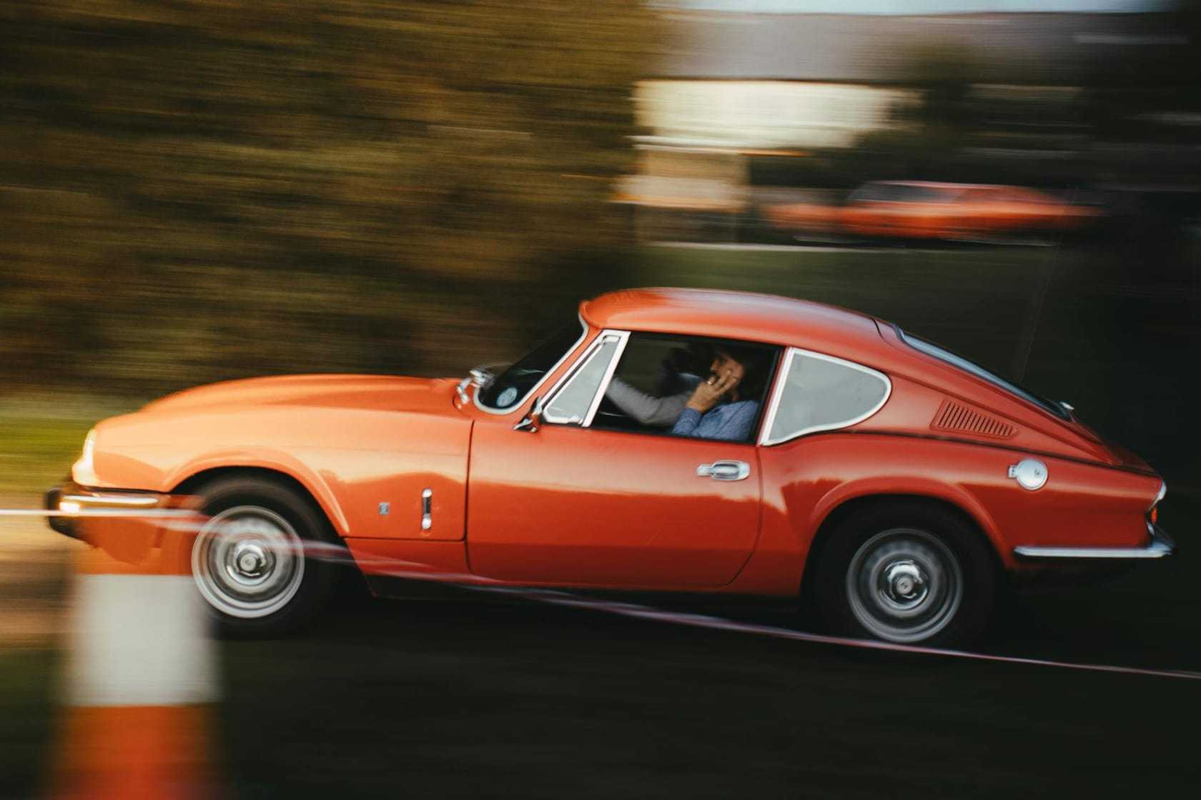 An orange Triumph GT6 fastback coupe photographed in motion on a road, side view