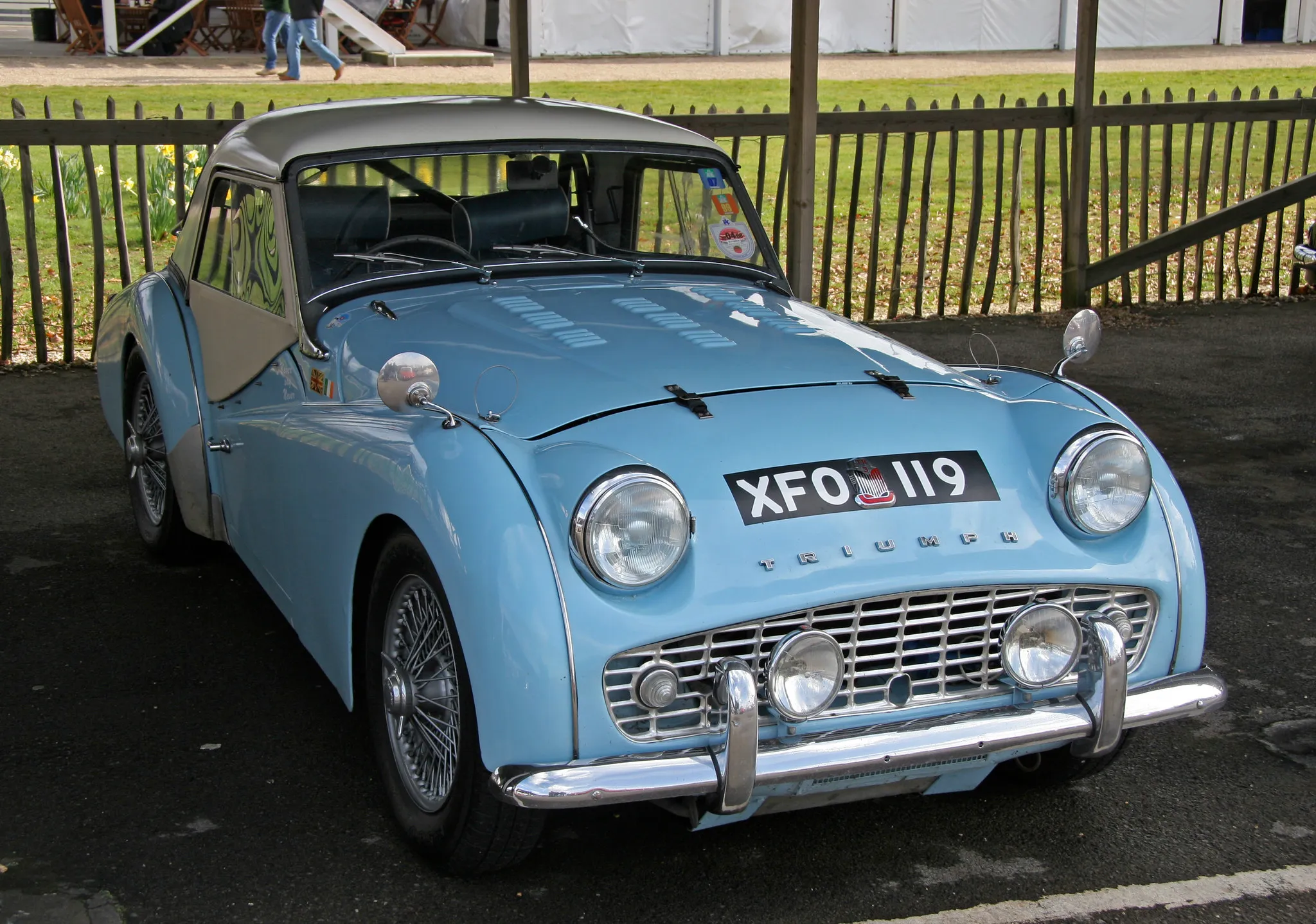 A pale blue Triumph TR3 with wire wheels and the hood raised, front three-quarter view