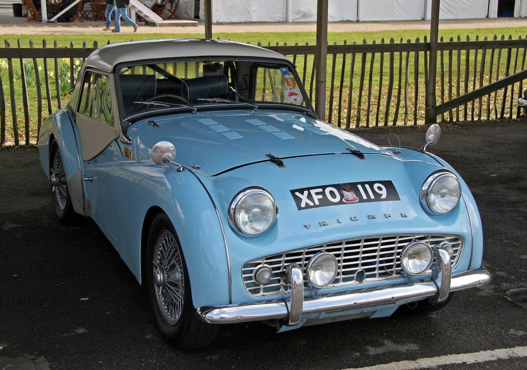 A pale blue Triumph TR3 with wire wheels and the hood raised, front three-quarter view