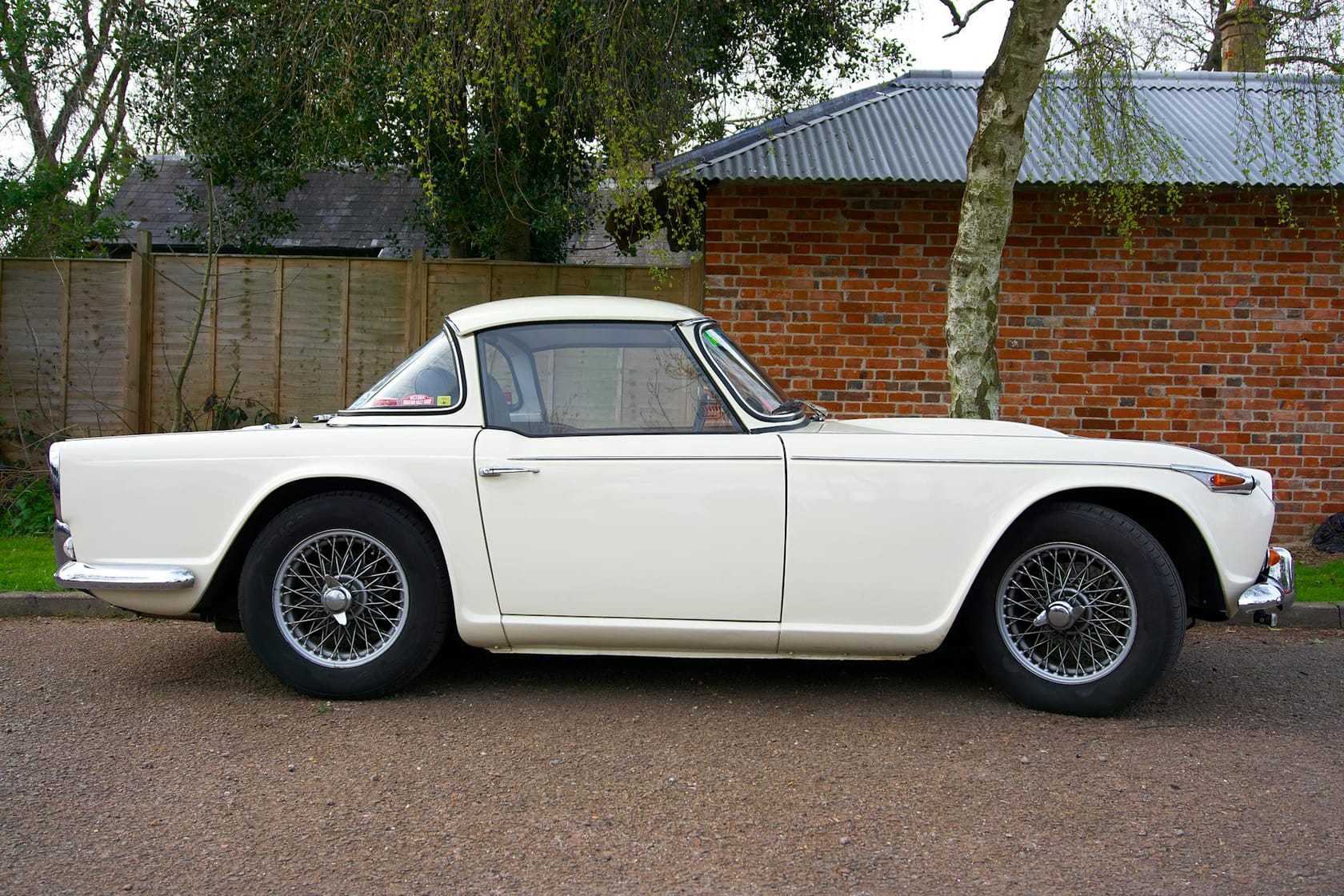 A white Triumph TR4 with its factory hardtop fitted, in side profile beside a brick barn