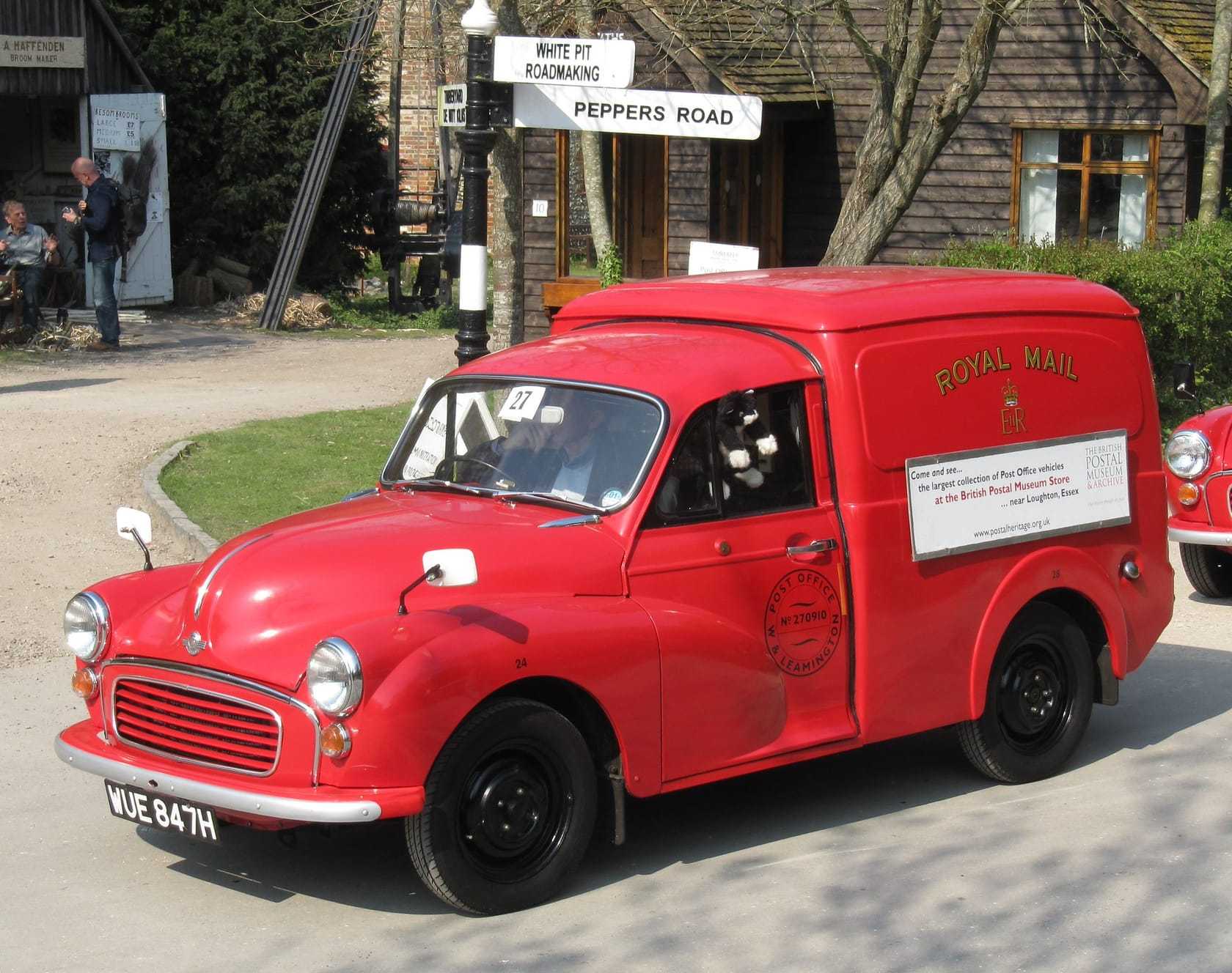 A red Royal Mail Morris Minor van, front three-quarter view, parked at a museum village on a sunny day