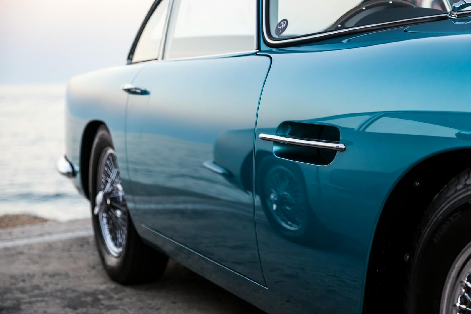 The flawless turquoise flank and chrome wire wheel of an Aston Martin DB5, parked beside calm water at dusk