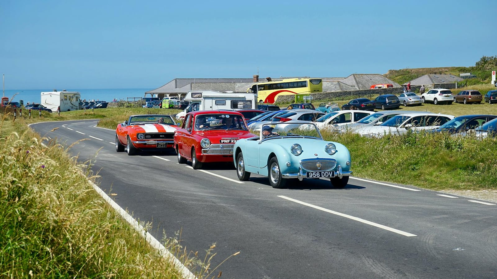 A light blue Austin-Healey Frogeye Sprite leading a line of classic cars on a coastal road