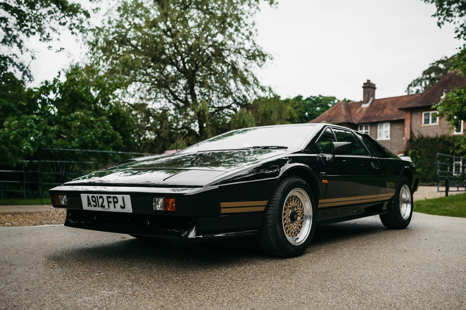 A black Lotus Esprit with a gold side stripe parked on a driveway in front of a country house