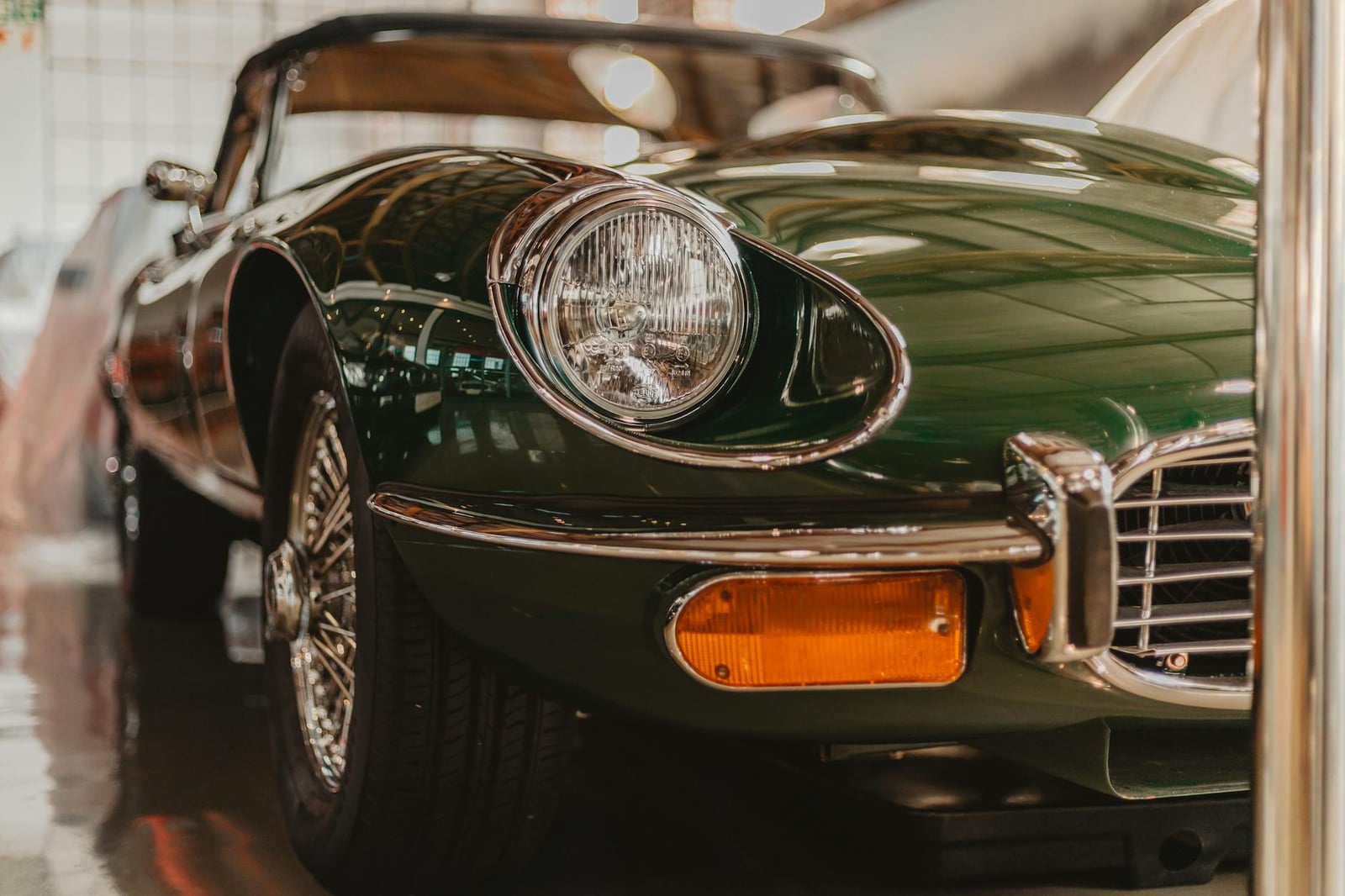 Low close-up of the open headlamp, chrome bumper and front wing of a dark green Jaguar E-Type