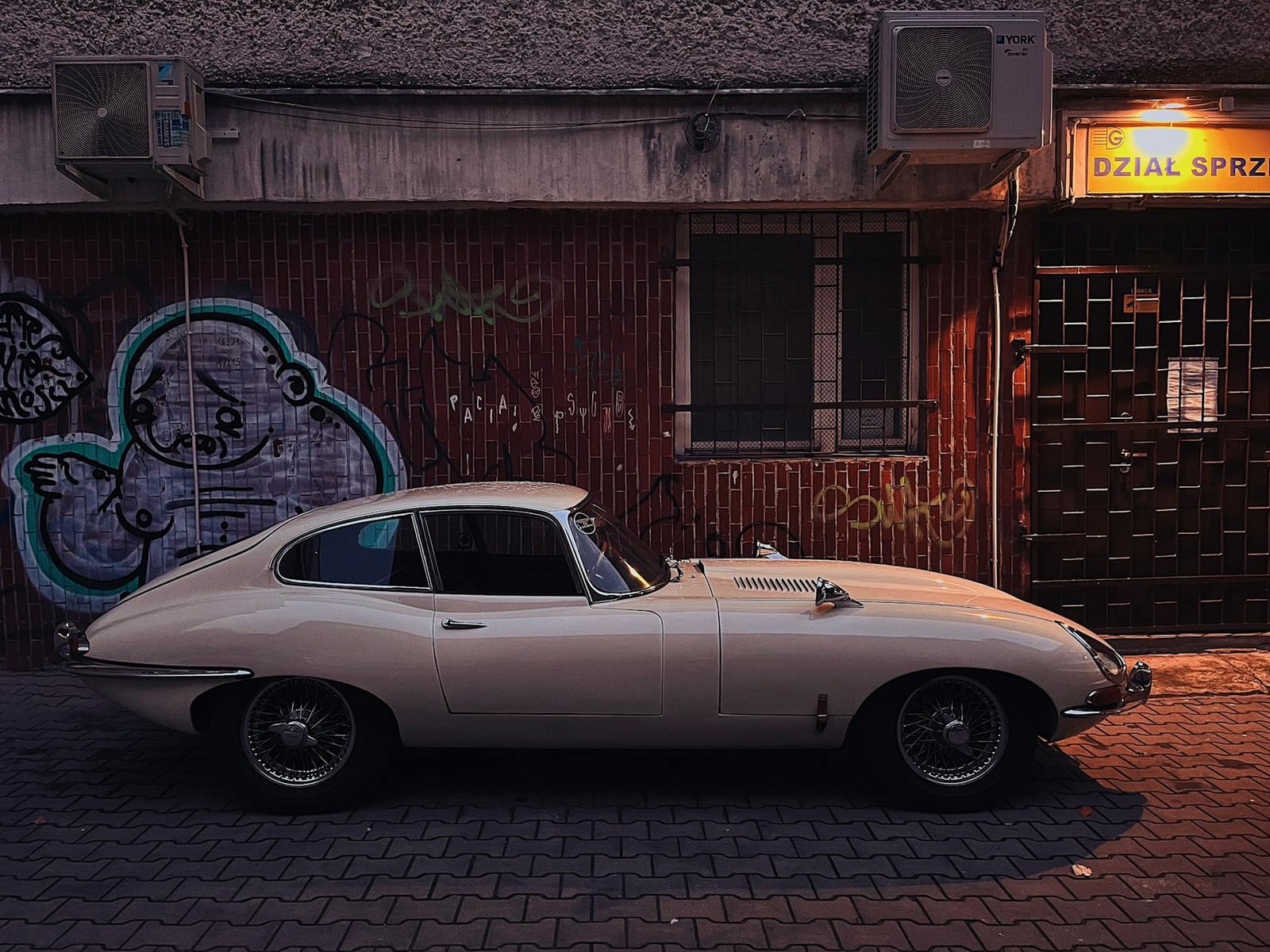 A cream Jaguar E-Type fixed-head coupe parked on a cobbled city street at dusk