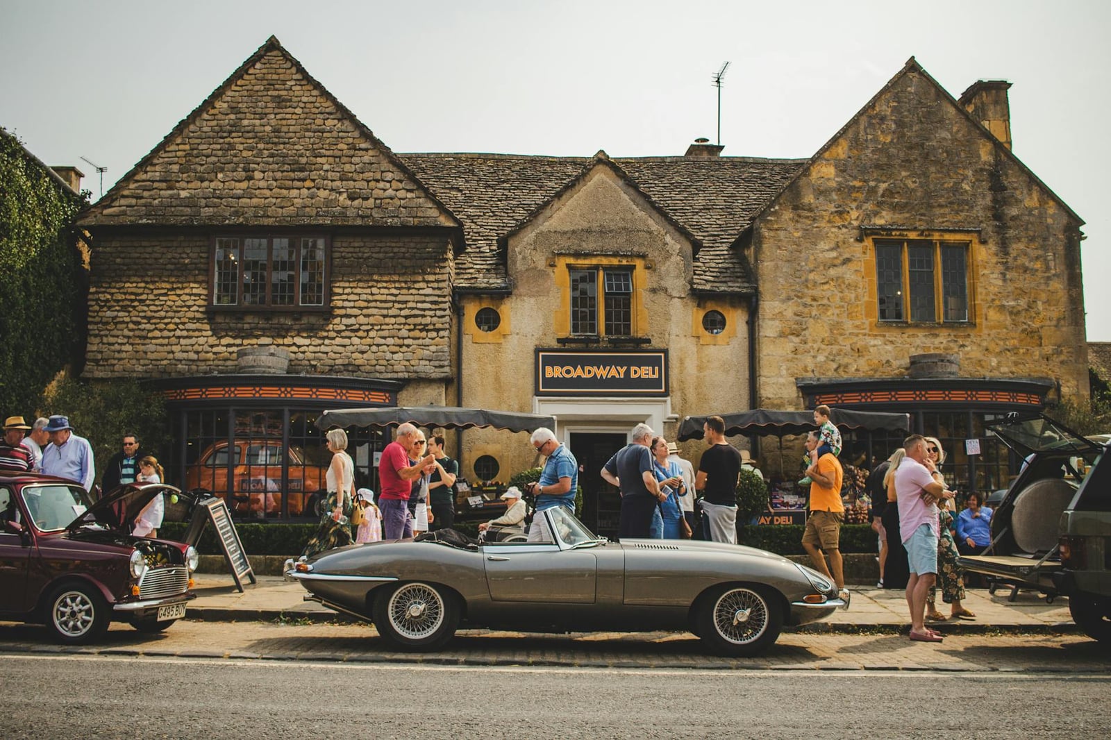 A Jaguar E-Type roadster parked in front of a honey-coloured stone building at a classic car gathering